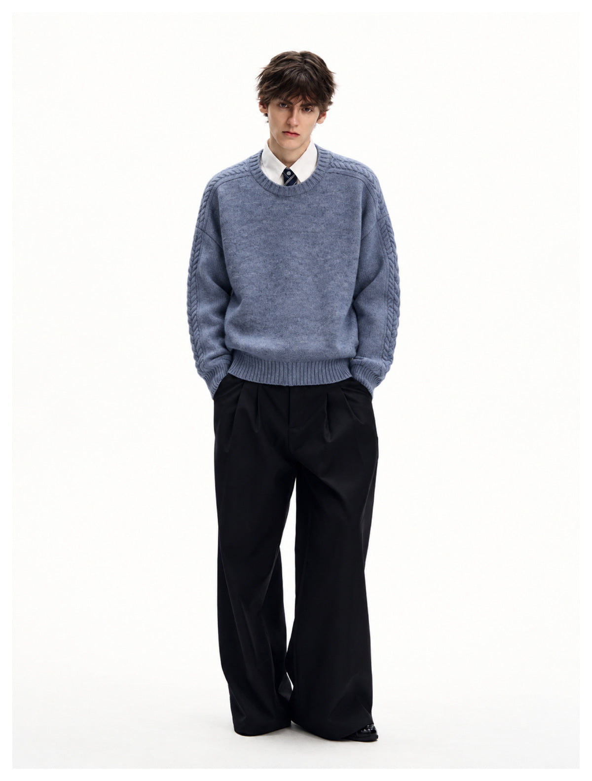 Full-length view of model in blue cable-knit sweater with white shirt, tie and wide black pleated trousers