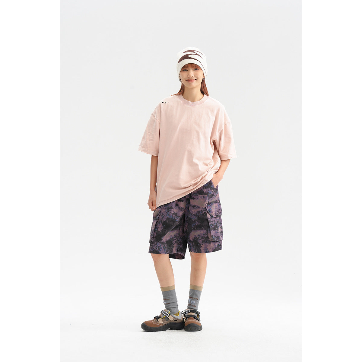 Woman wearing an oversized pale pink T-shirt and purple camouflage cargo shorts against a white studio background.