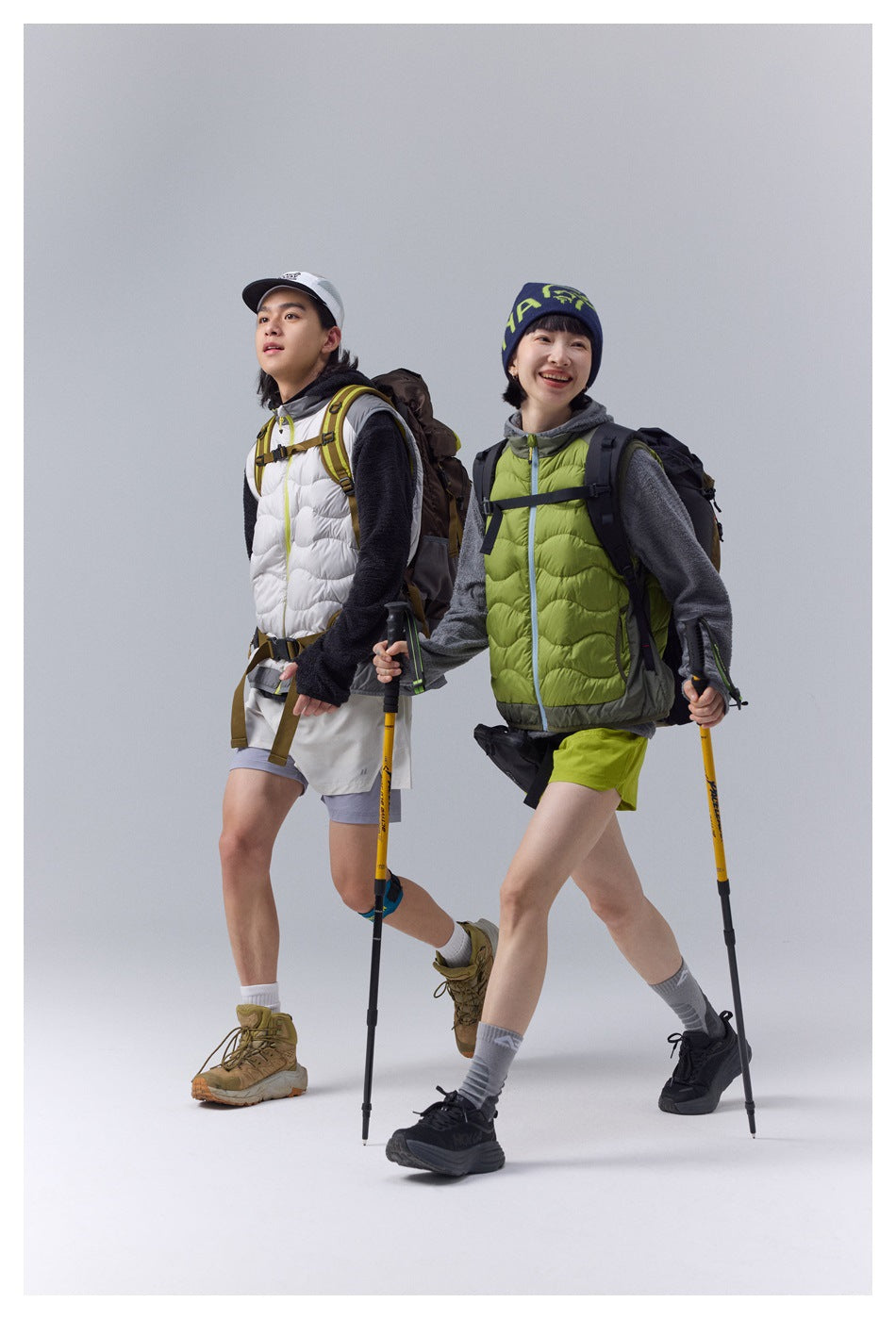 Two hikers wear white and lime-green quilted vests over dark fleece layers, with shorts, backpacks and trekking poles.
