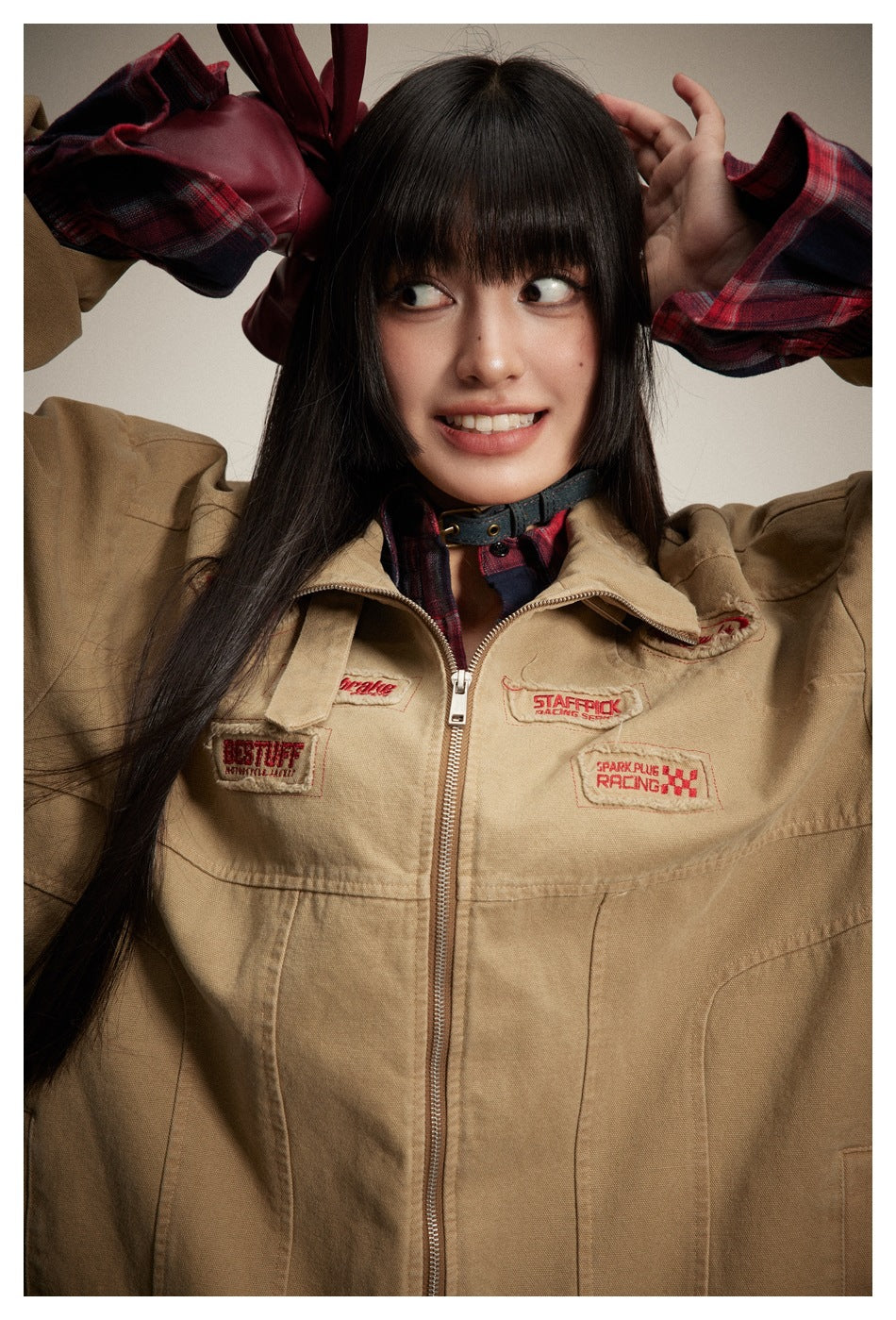 Close view of a model in a tan zip jacket with embroidered patches and a red-and-navy plaid shirt.
