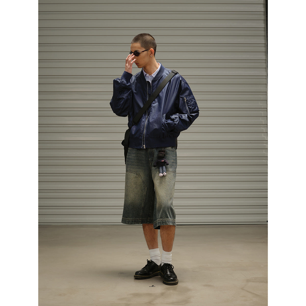 Model wearing a navy zip-front jacket and oversized faded denim shorts, with a crossbody bag and small plush charm.