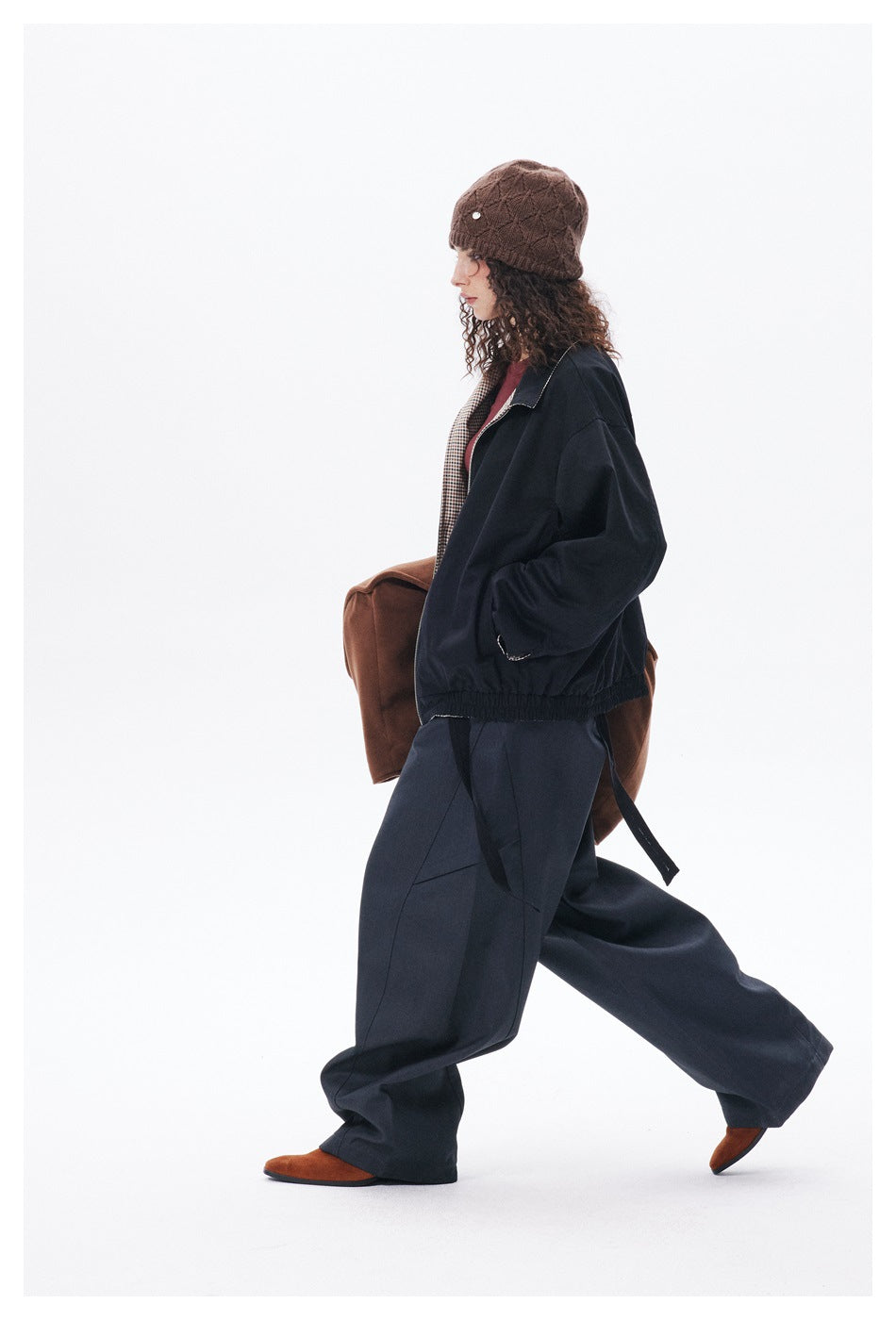 Side view of a model walking in a black jacket with checked lining and loose dark trousers, wearing a brown knit beanie.
