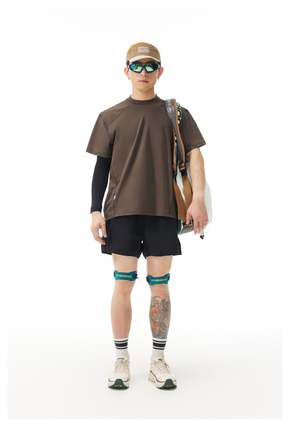 Model wearing brown short-sleeve T-shirt with black shorts, black arm sleeve, sunglasses, cap and racquet bag