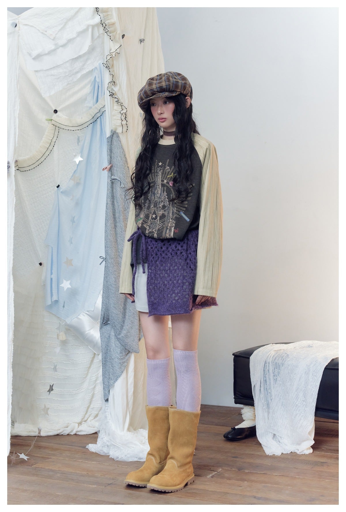 Model in the castle-print raglan sweatshirt, turned slightly to the side, with a plaid cap and purple knit skirt
