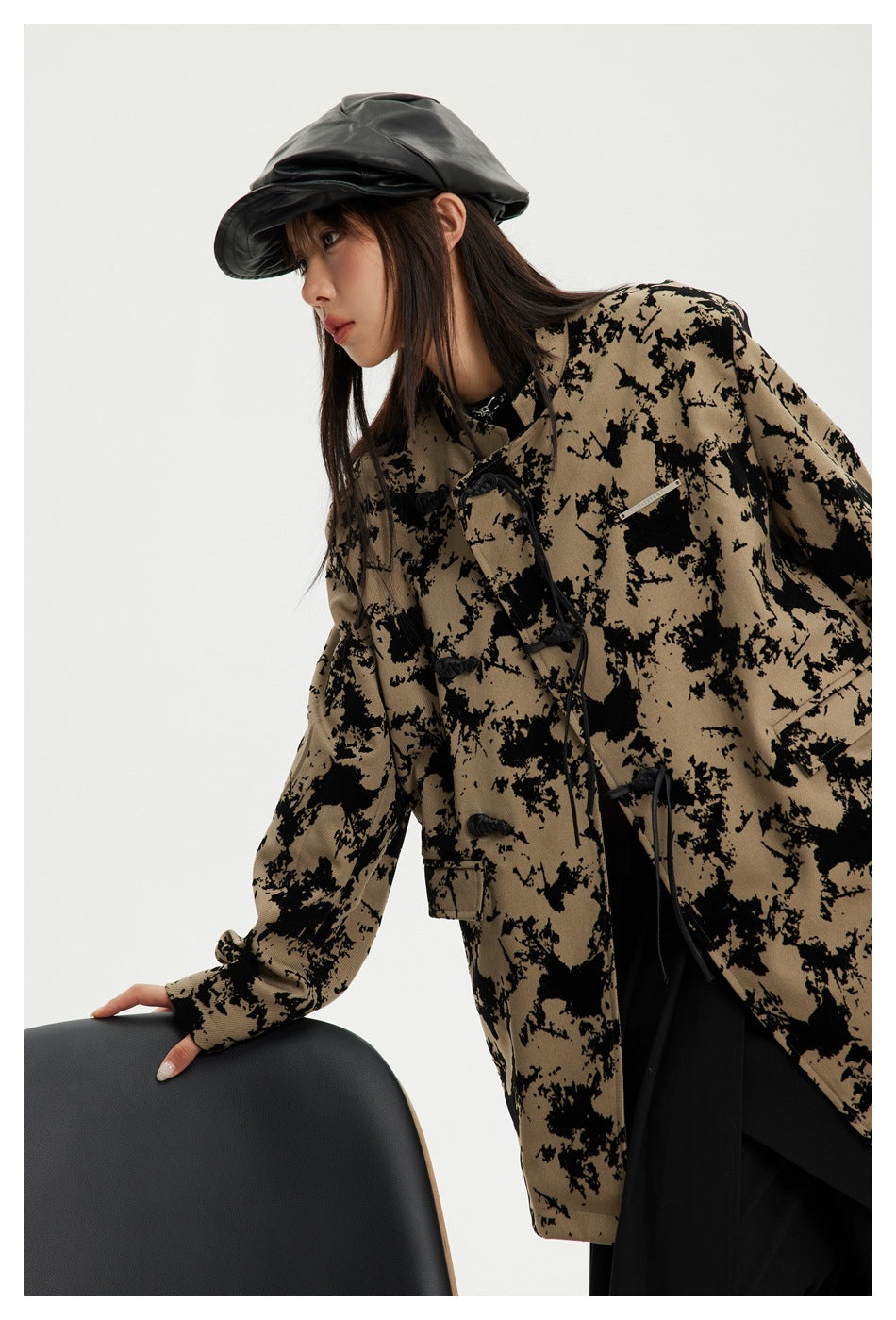 Close view of a model in a tan jacket with an irregular black pattern and toggle closures, leaning on a chair.