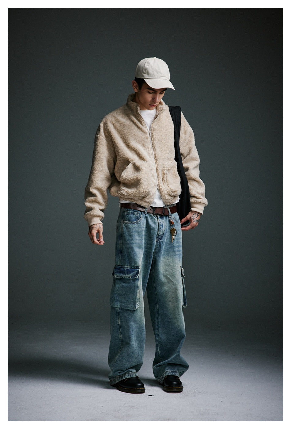 Full-length model wearing a beige zip-up fleece jacket with front pockets, loose blue cargo jeans, and a white cap.