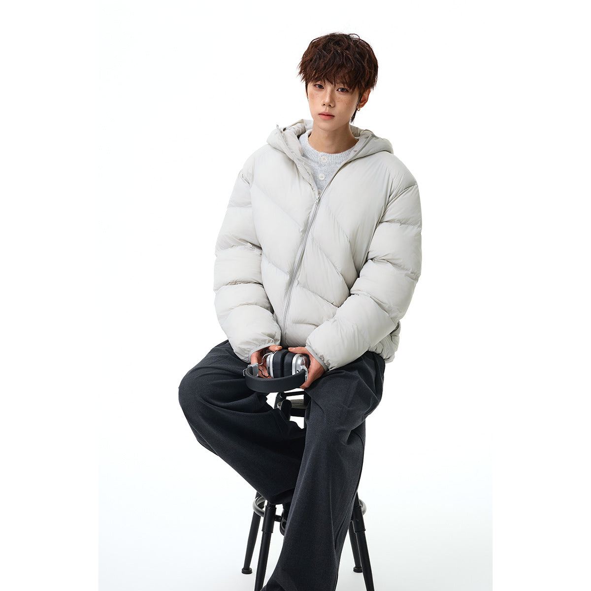 Seated model wearing a light gray hooded puffer jacket, shown from the front with the zipper partly open.