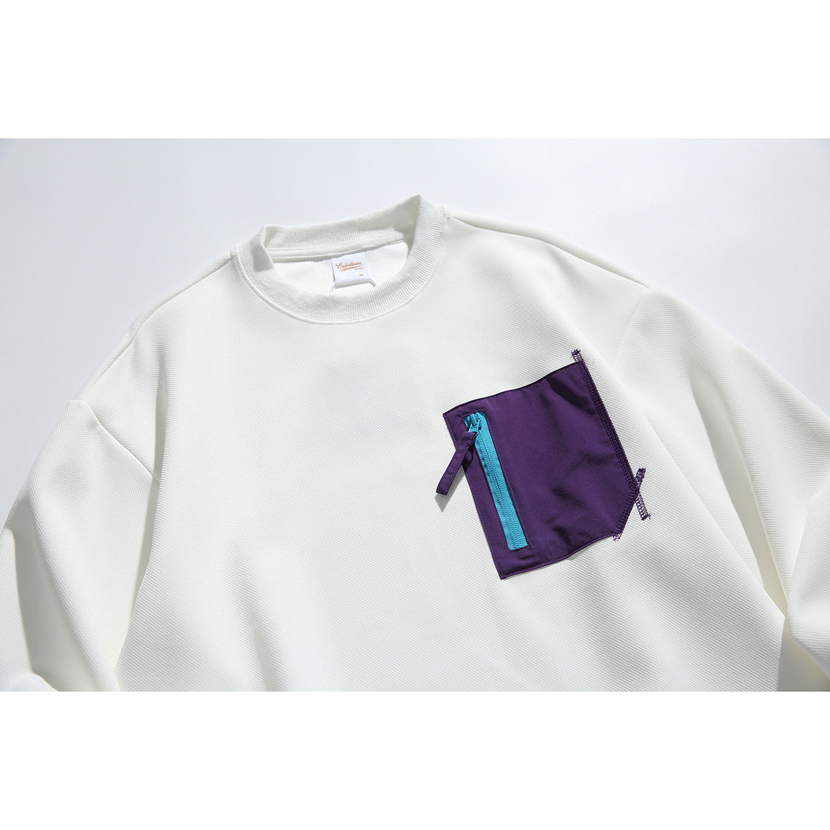 Close view of an ivory crewneck sweatshirt with a purple chest pocket, bright cyan zipper, and purple pull tab.