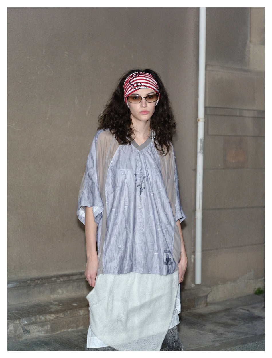 Model wearing a pale gray V-neck top with sheer taupe sleeves over a white layered skirt.