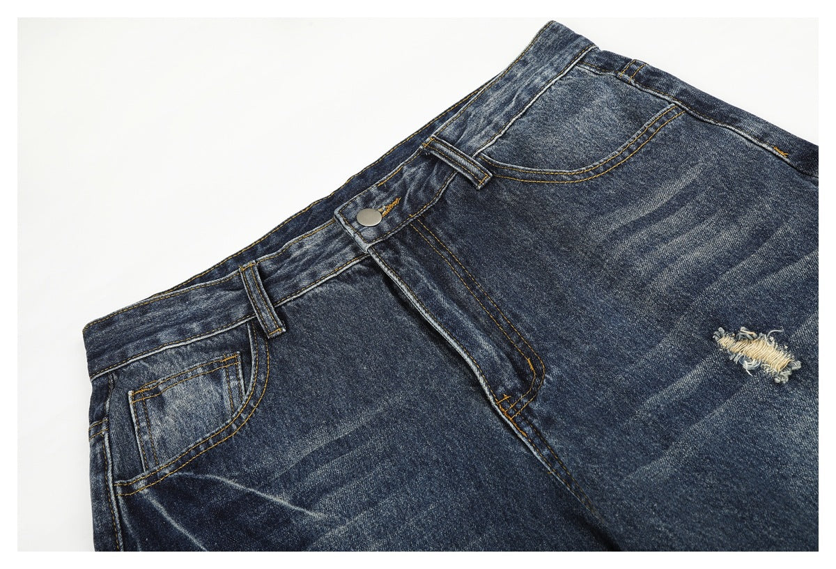 Angled close-up of dark blue jeans with a buttoned waistband, front pocket, golden stitching, and a frayed tear.
