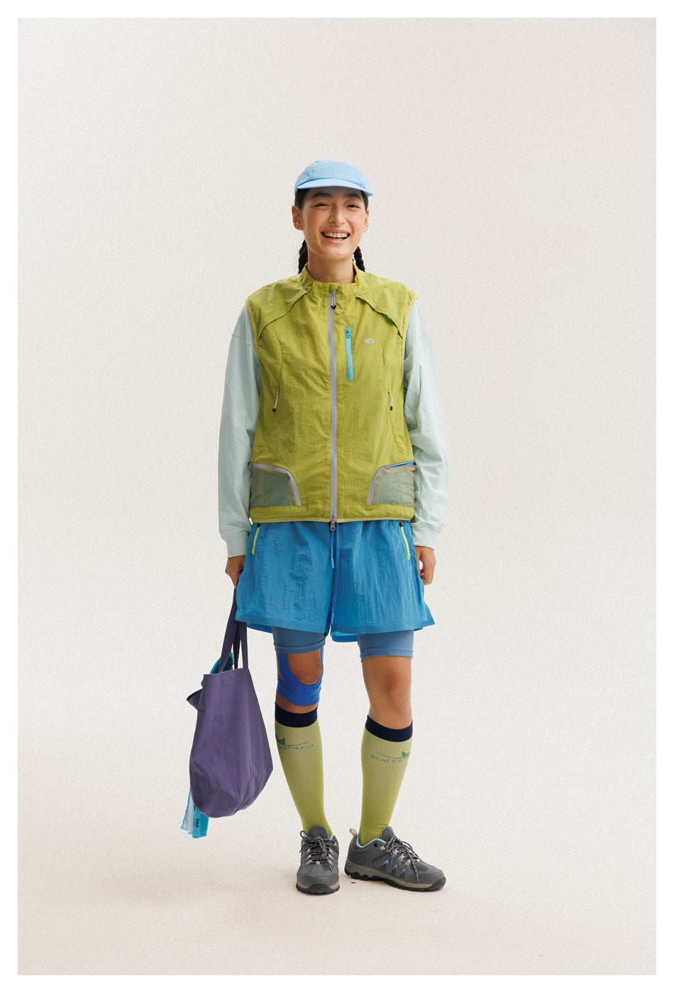 Full-length model wearing a lime-green sleeveless zip vest, pale mint top, and bright blue athletic shorts.