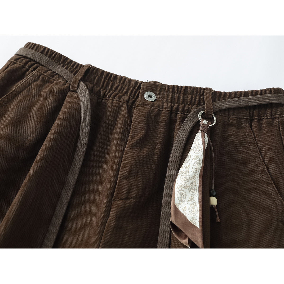 Close-up of brown trousers with an elastic waist, drawcords, metal button, and patterned hanging accent.