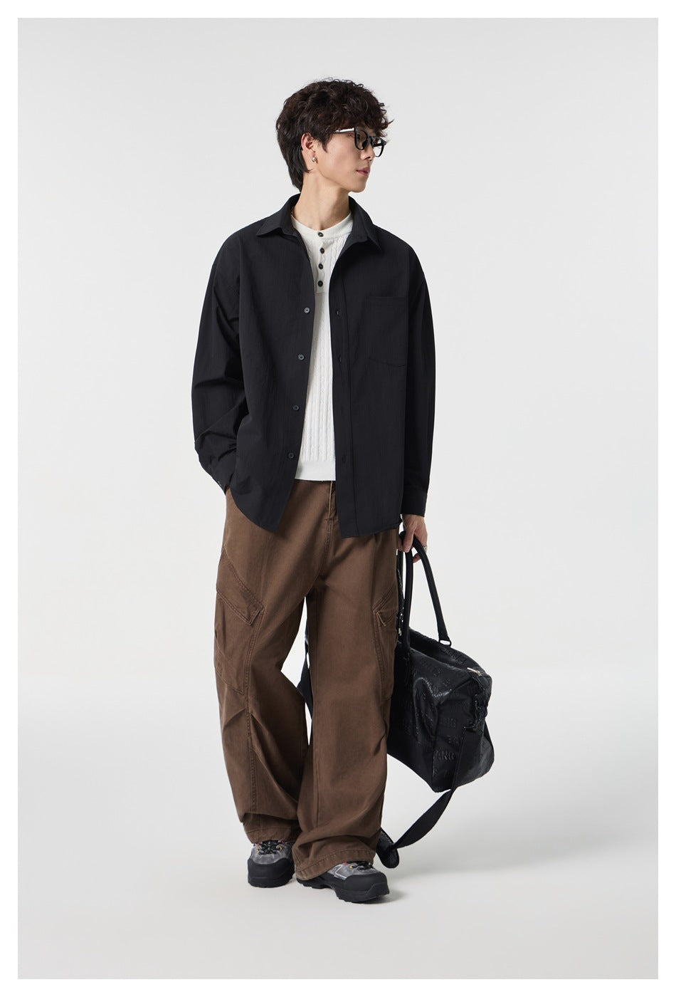 Model wearing a black button-front shirt open over a white knit top with brown cargo pants and holding a black bag