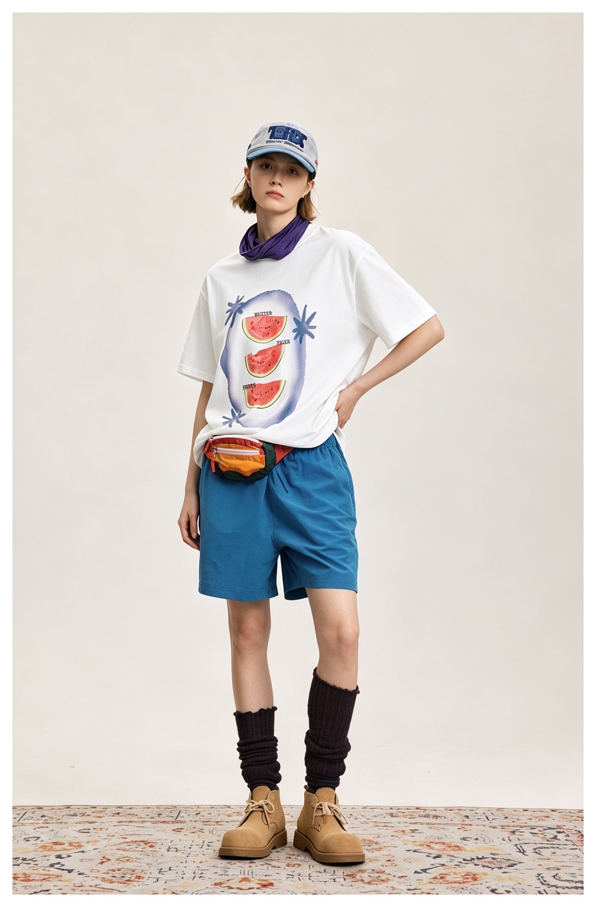 Person wearing the white watermelon-print T-shirt with blue shorts, a cap, a scarf, a colorful waist bag and tan boots