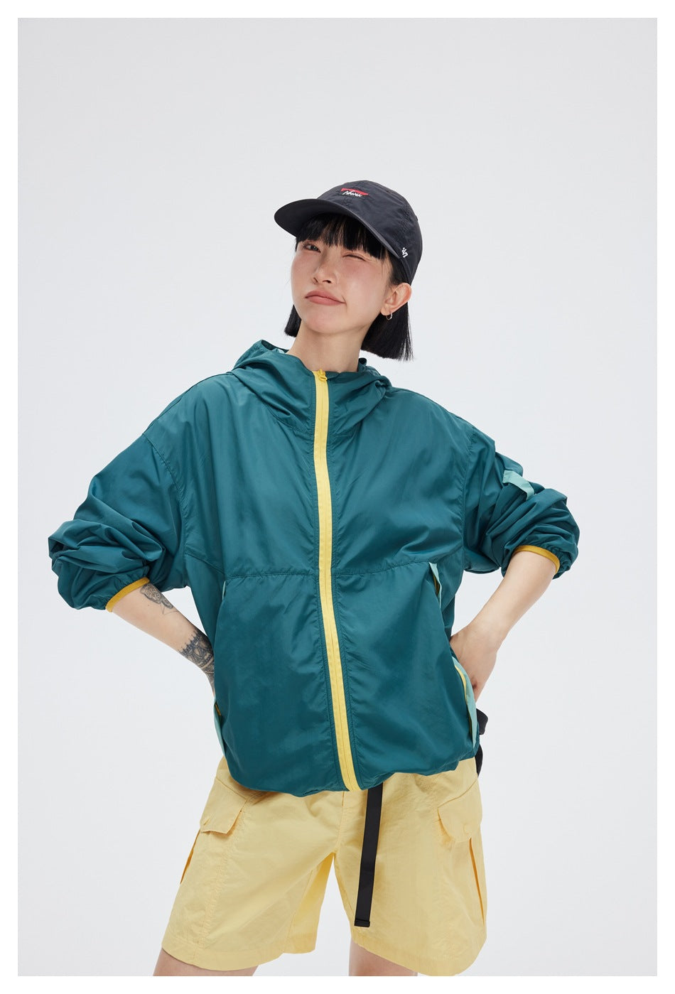 Model wearing a teal hooded jacket with a yellow zipper and cuffs, posed with hands on hips.