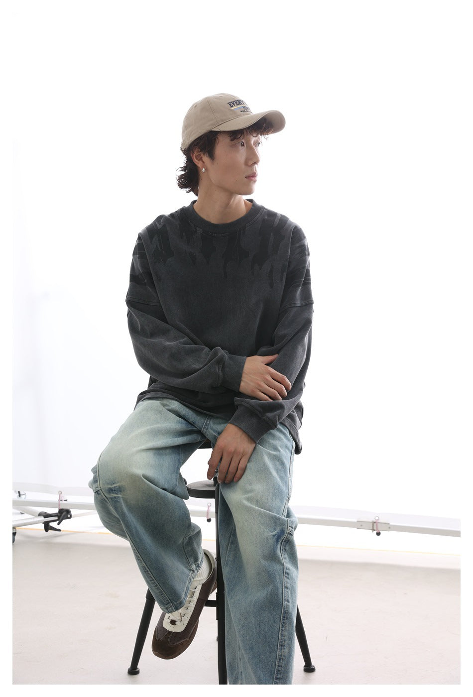 Seated model wearing a charcoal crewneck sweatshirt with dark shoulder graphics and faded blue jeans.
