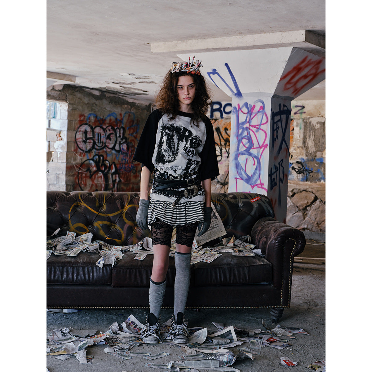 Model wears a black-sleeved graphic raglan shirt with a striped skirt and layered belts in a graffiti-covered room.