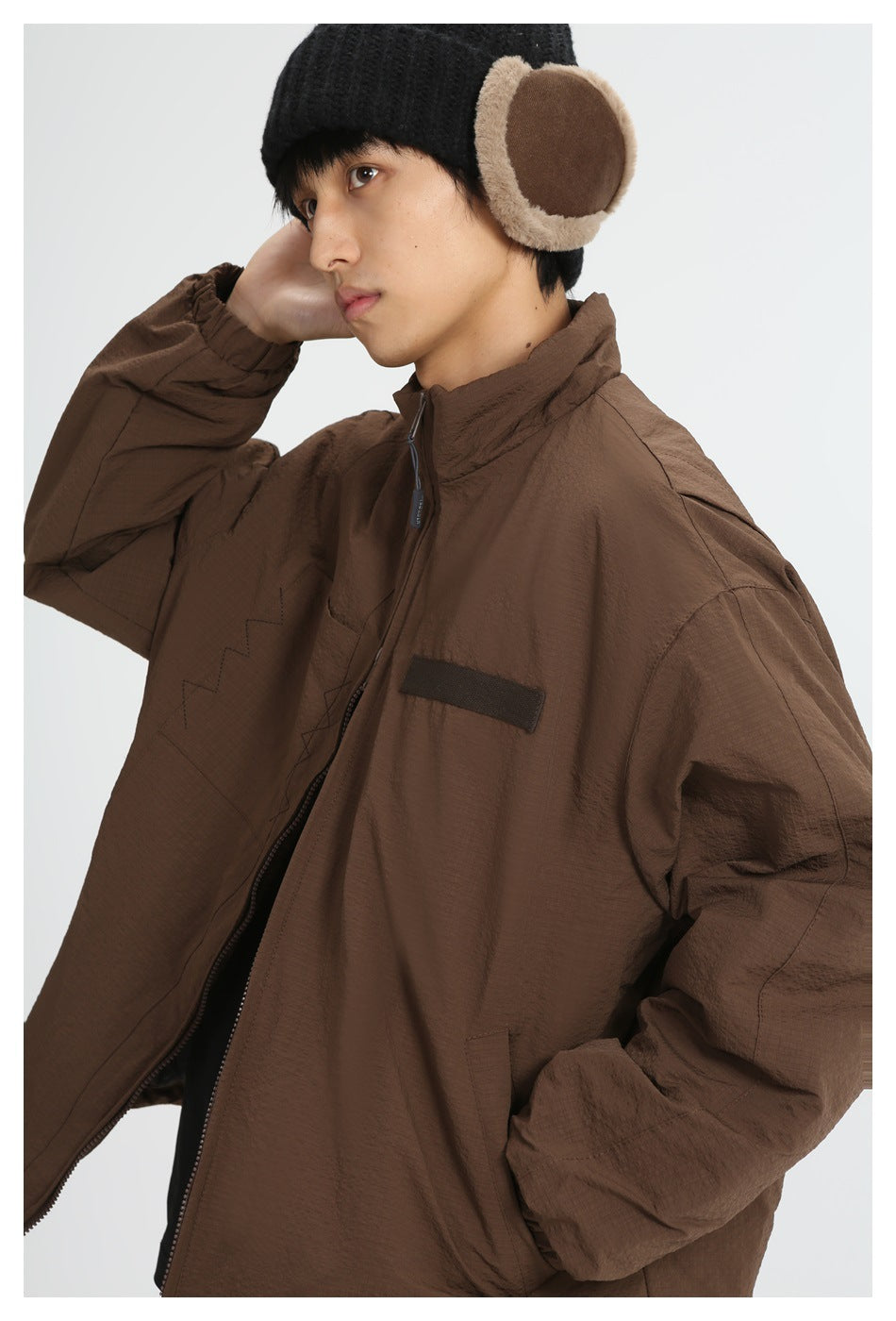 Model wearing a brown zip-front jacket with a high collar and elastic cuffs, paired with a black knit earflap hat.