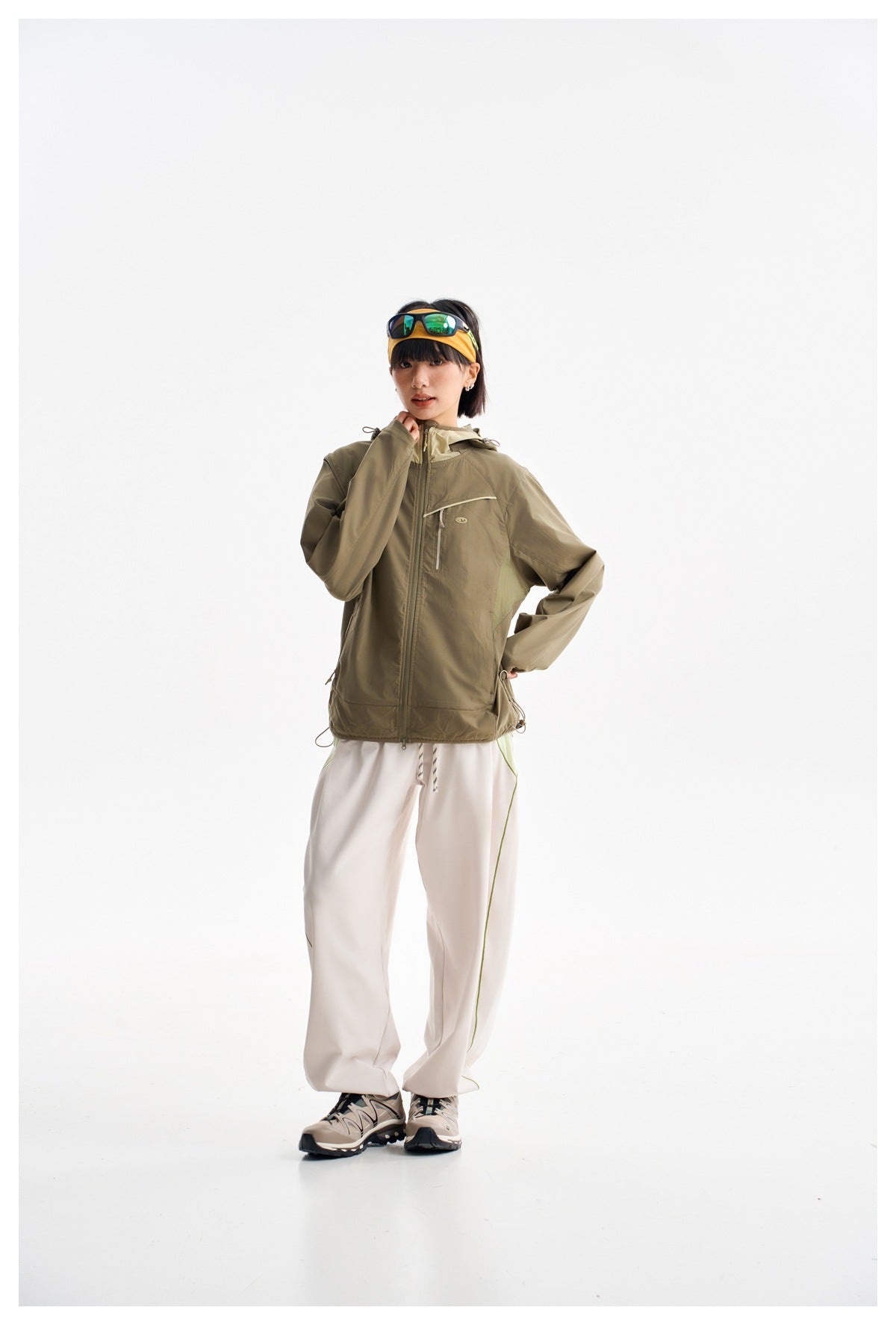Model wearing a zipped olive hooded jacket and loose light pants, shown from the front.