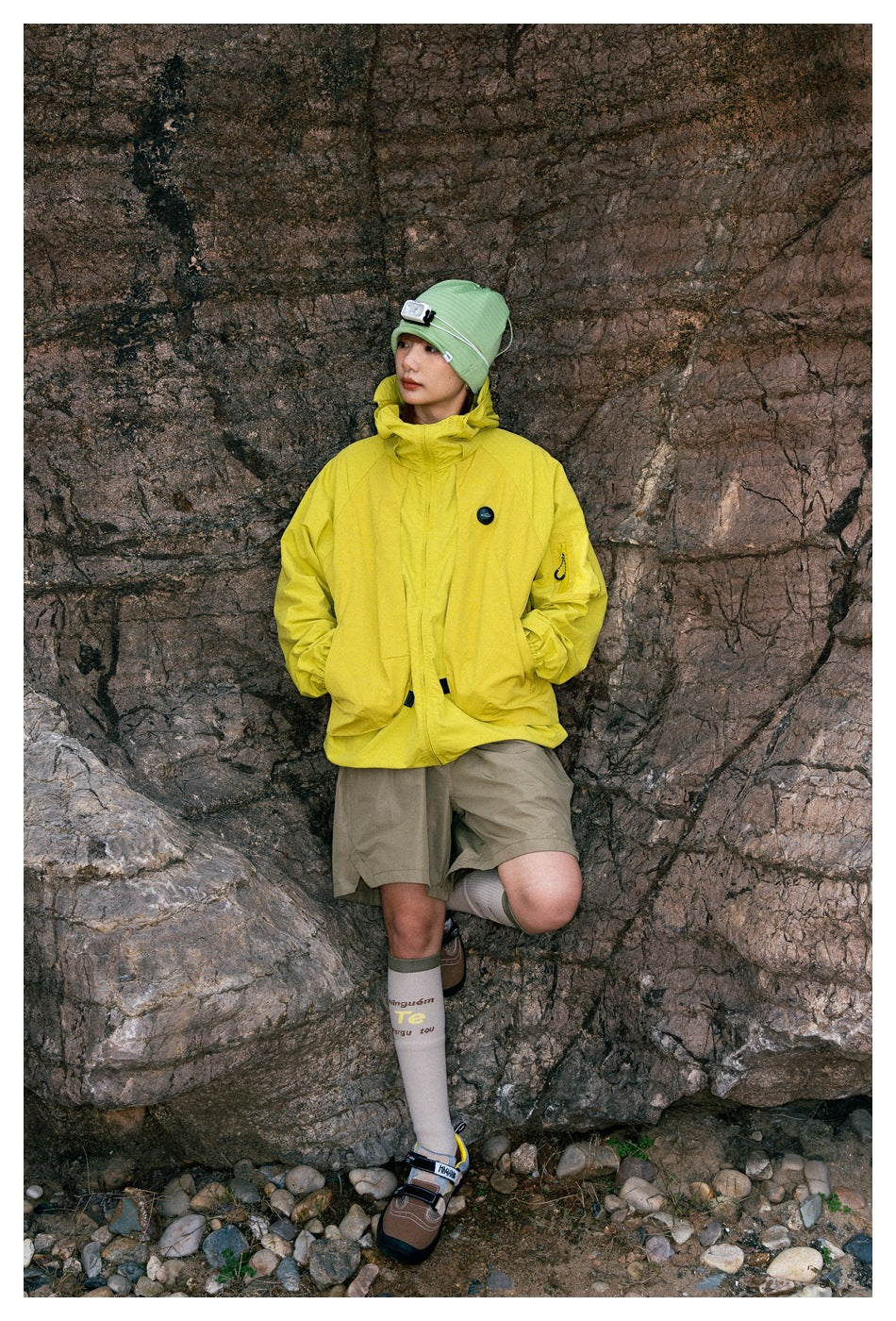 Model wearing a bright yellow hooded jacket with large pockets and taupe shorts, posed against a rocky backdrop.