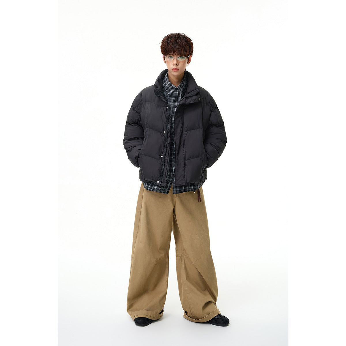 Model wearing a black quilted puffer jacket over a plaid shirt and wide tan trousers.