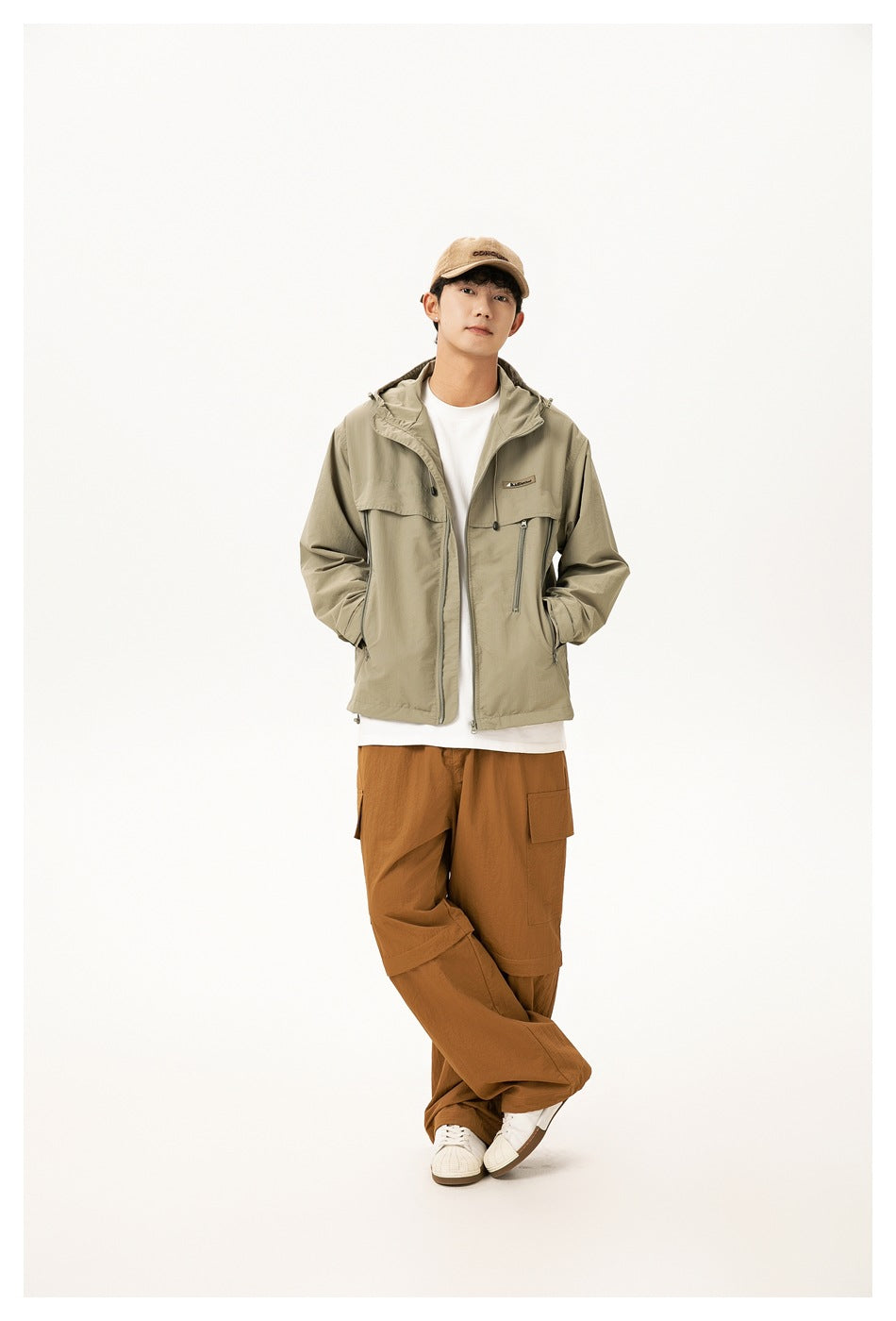 Model wearing light green hooded zip jacket over white tee with brown cargo pants and cap, hands in pockets
