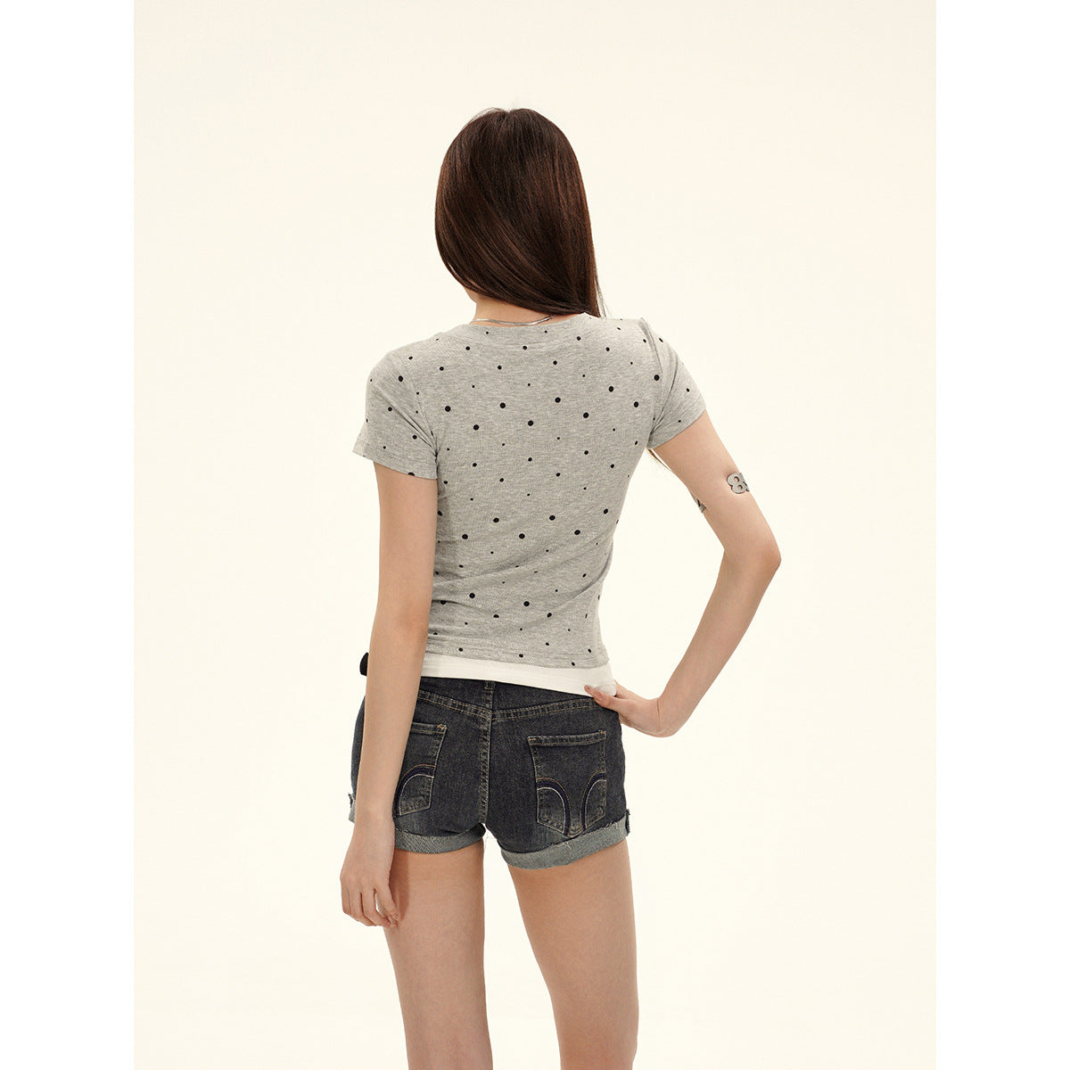 Back view of a model wearing a light gray tee with scattered black dots and dark cuffed denim shorts.