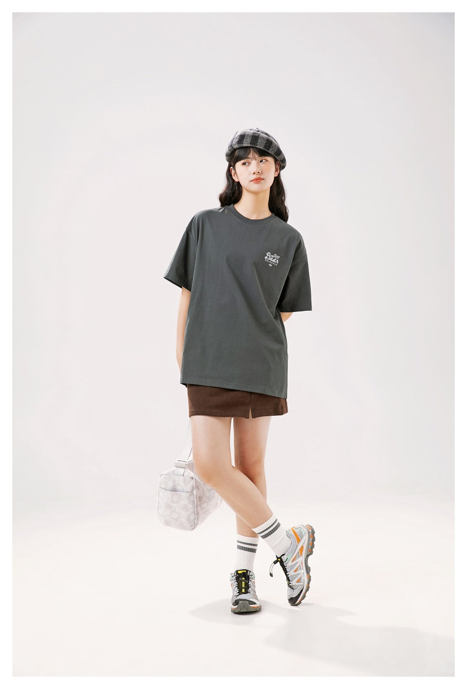 Model wearing an oversized charcoal gray T-shirt with a small white chest print, brown shorts, and a checked cap.