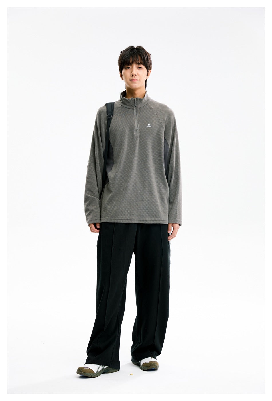 Model standing in a gray half-zip pullover with black wide-leg pants and a backpack strap, front view