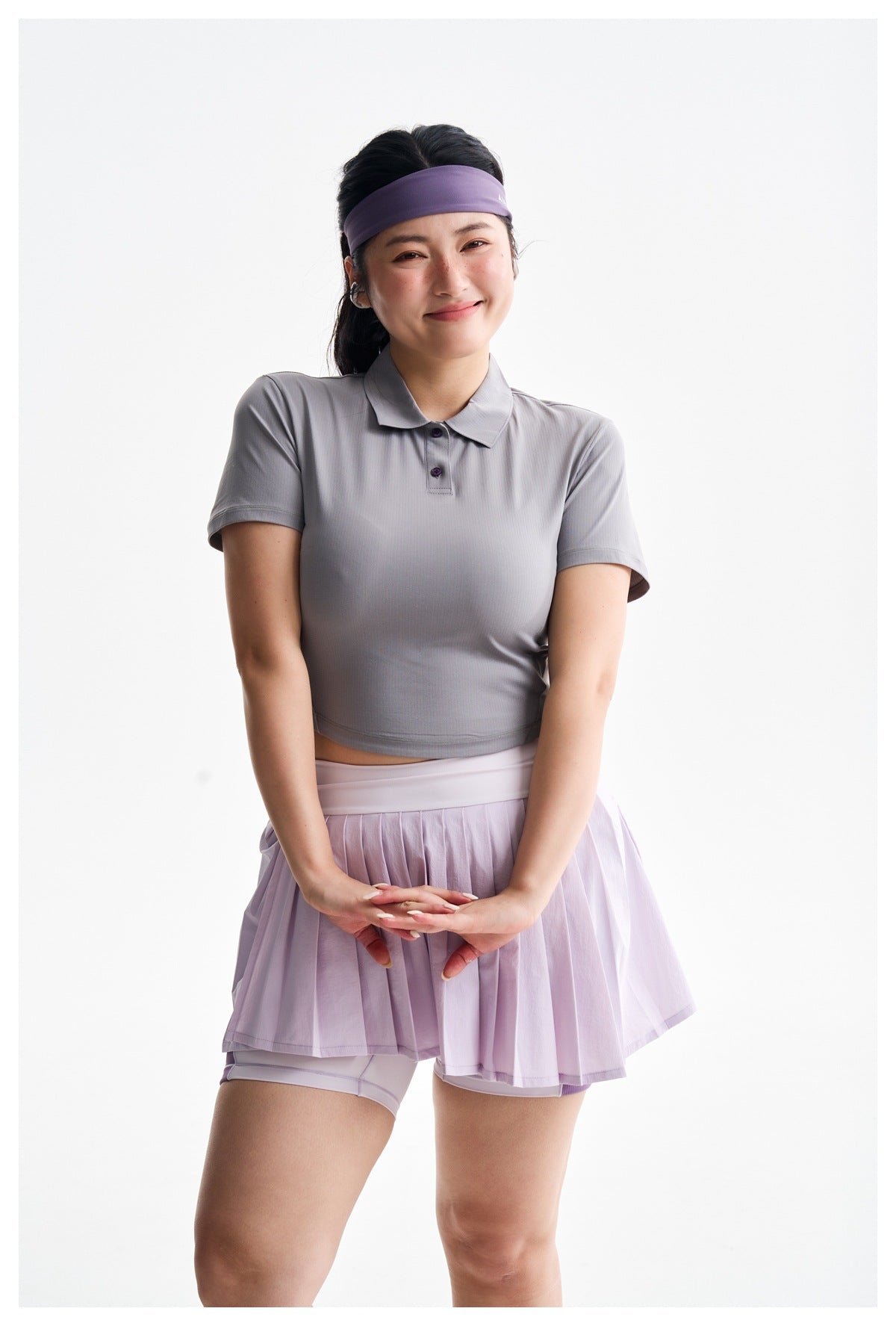 A woman models a gray cropped polo and lilac pleated skort with fitted undershorts against a white background.