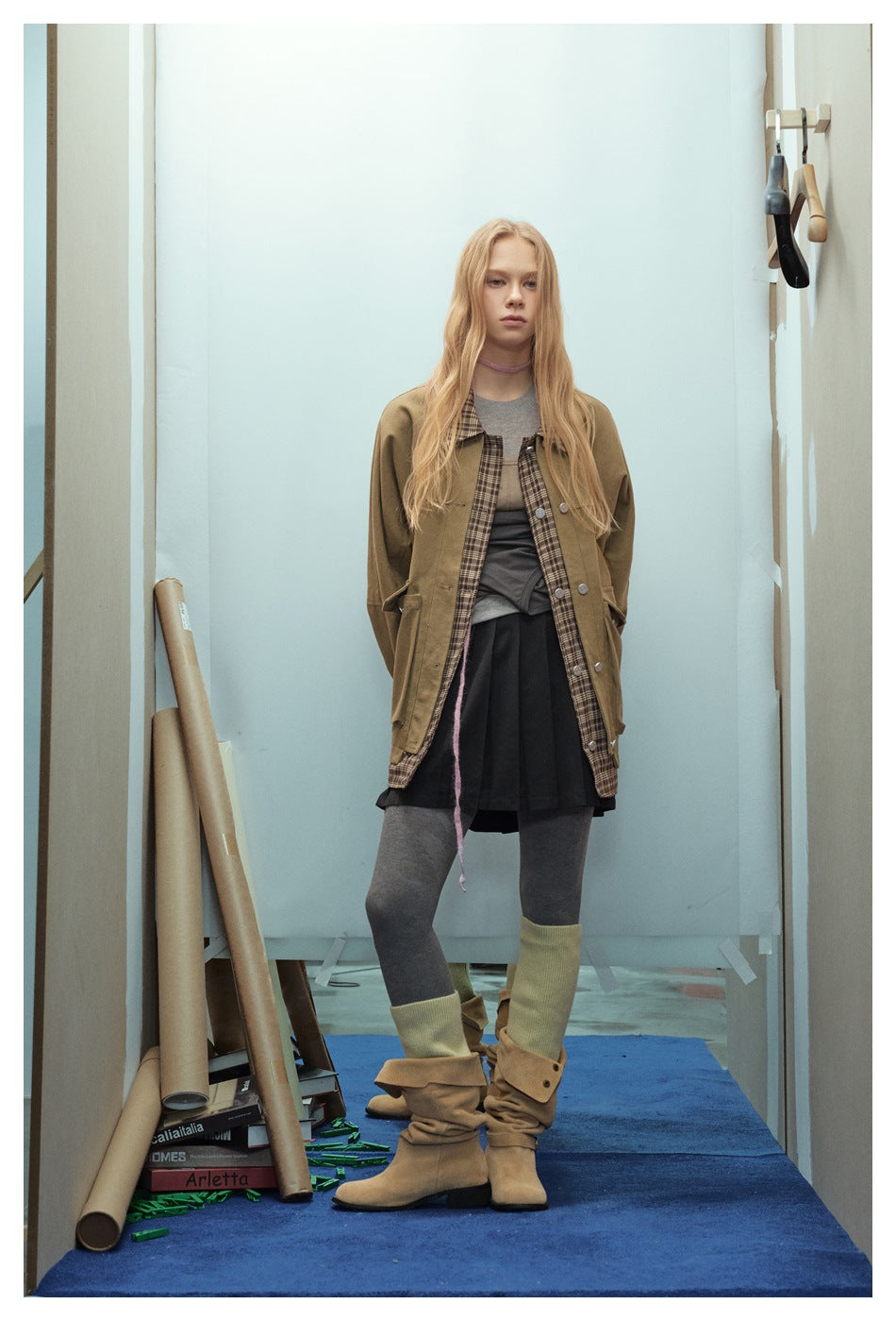 Model standing in a tan jacket with plaid trim, gray layered top, black pleated skirt, gray tights, and tan boots.