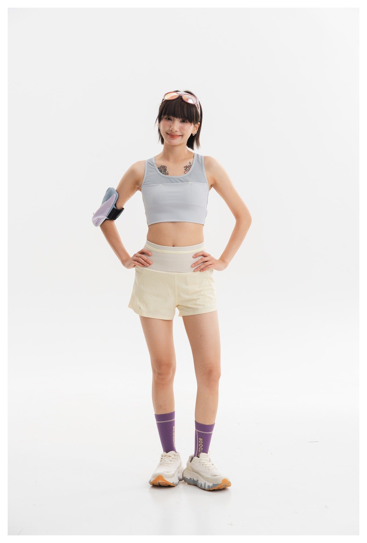 Model wearing a light gray cropped athletic tank and pale yellow shorts, with a padded arm accessory and purple socks.