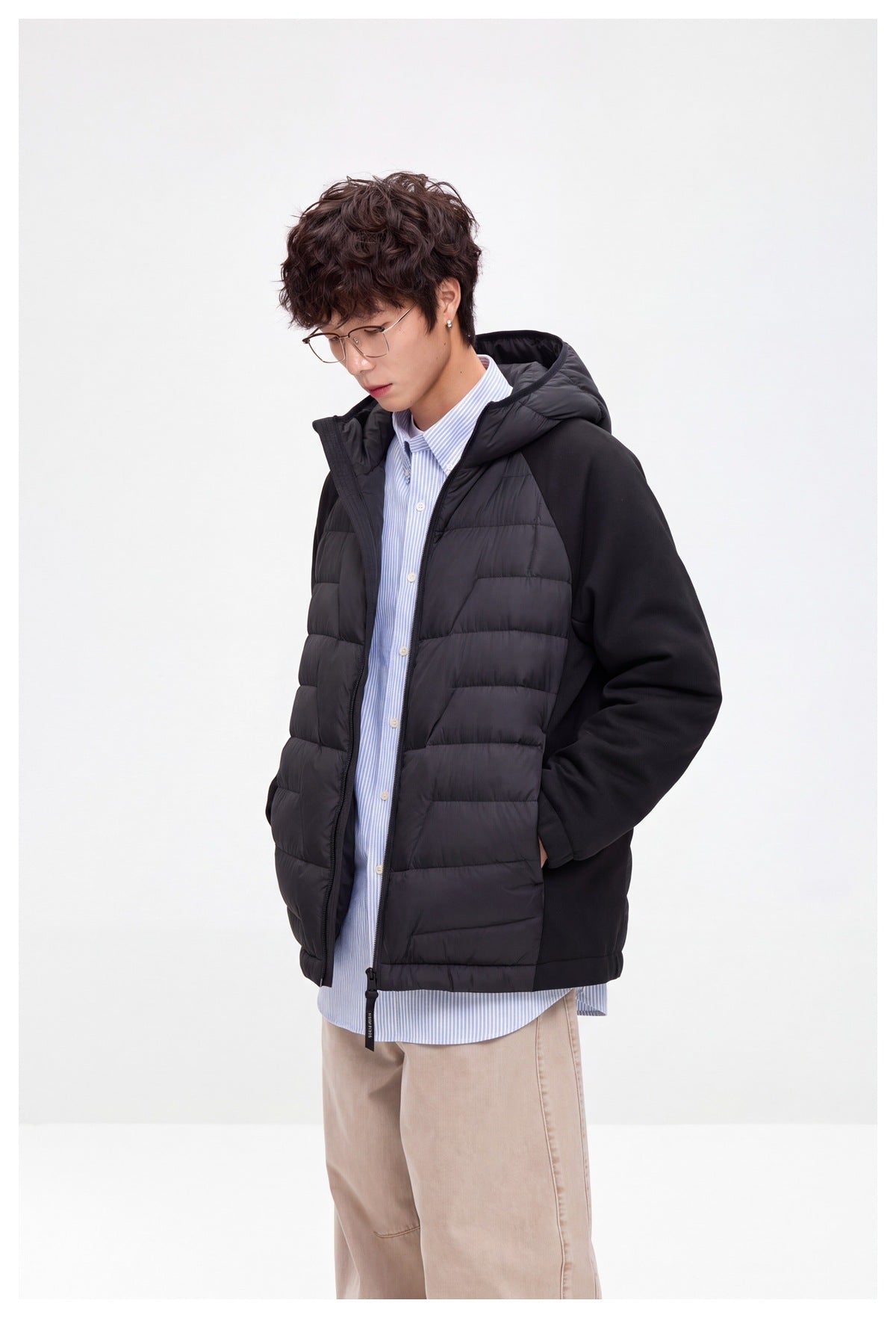 Man in black hooded quilted jacket with knit sleeves worn open over a blue striped shirt, hand in pocket