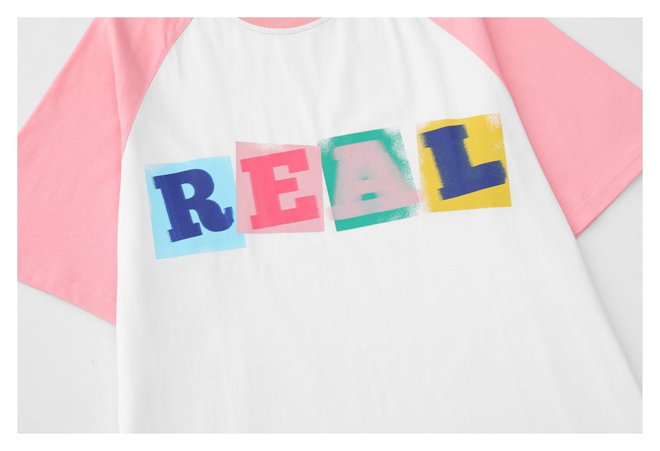 Close-up of a white T-shirt with pink raglan sleeves and colorful block-letter “REAL” print across the chest.