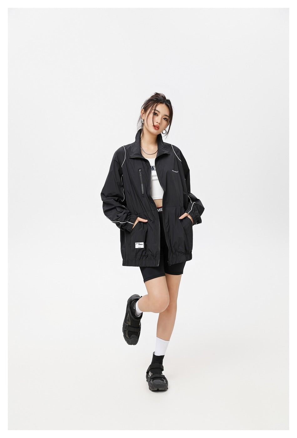Woman wearing a black oversized full-zip stand-collar jacket with white piping over a white top and black shorts