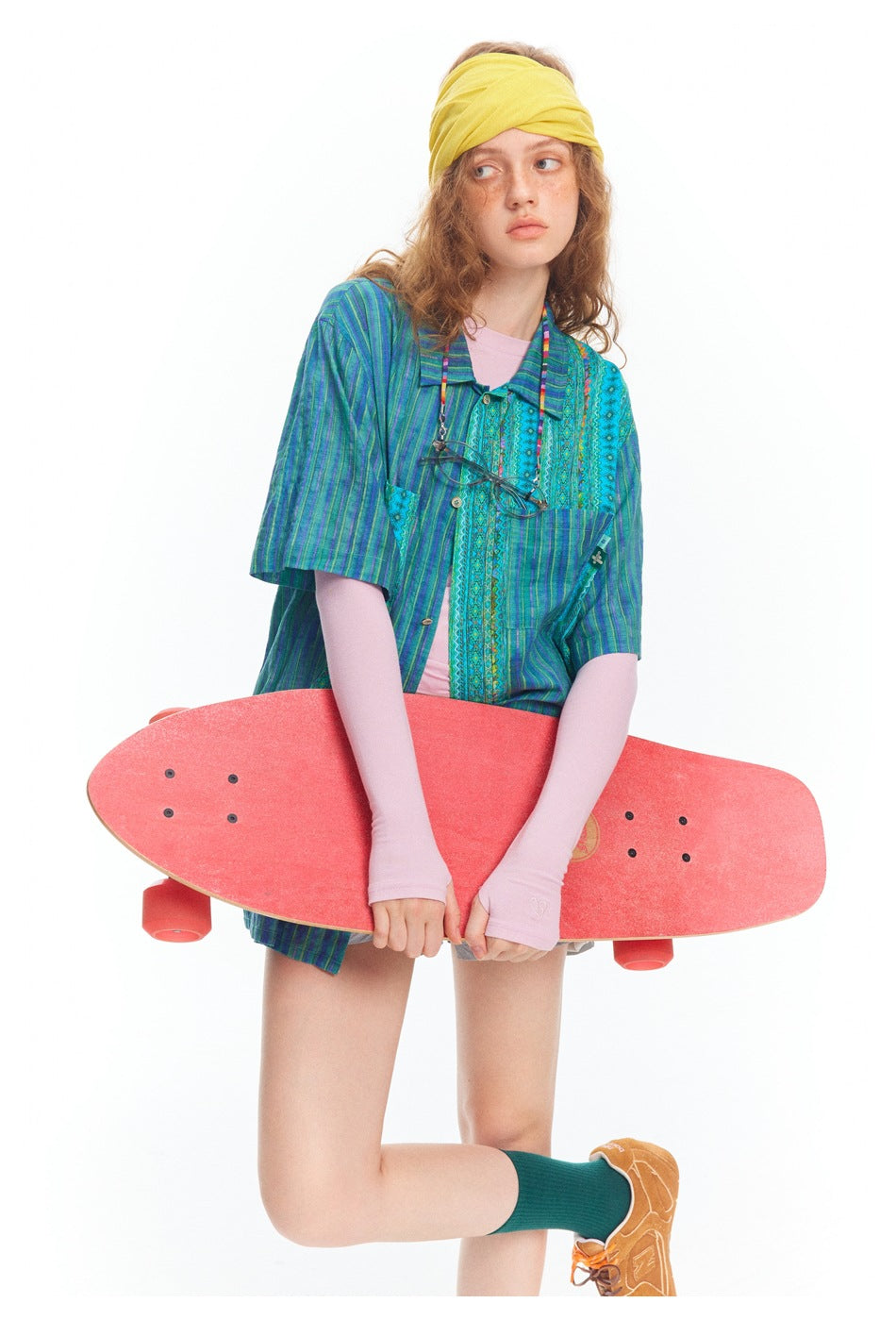 Model wearing a blue-green patterned shirt over pink sleeves and holding a red skateboard across the front.