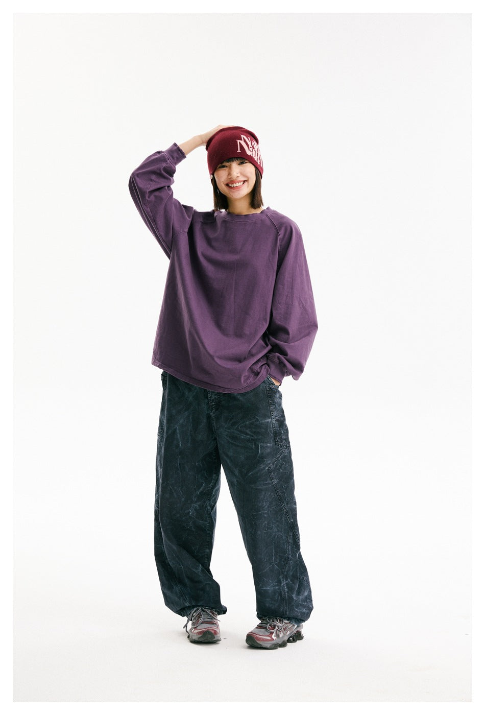 Smiling model wearing a purple oversized raglan sweatshirt, dark patterned pants, and a burgundy beanie with white lettering.