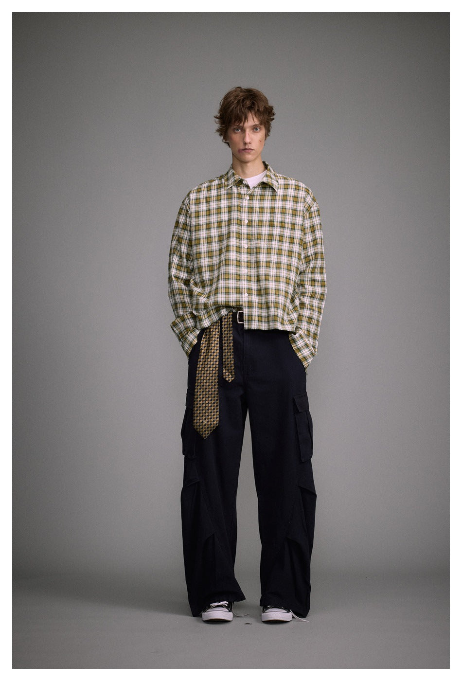 Full-length model in a white, green, and mustard plaid shirt, black cargo pants, and a patterned tie at the waist.