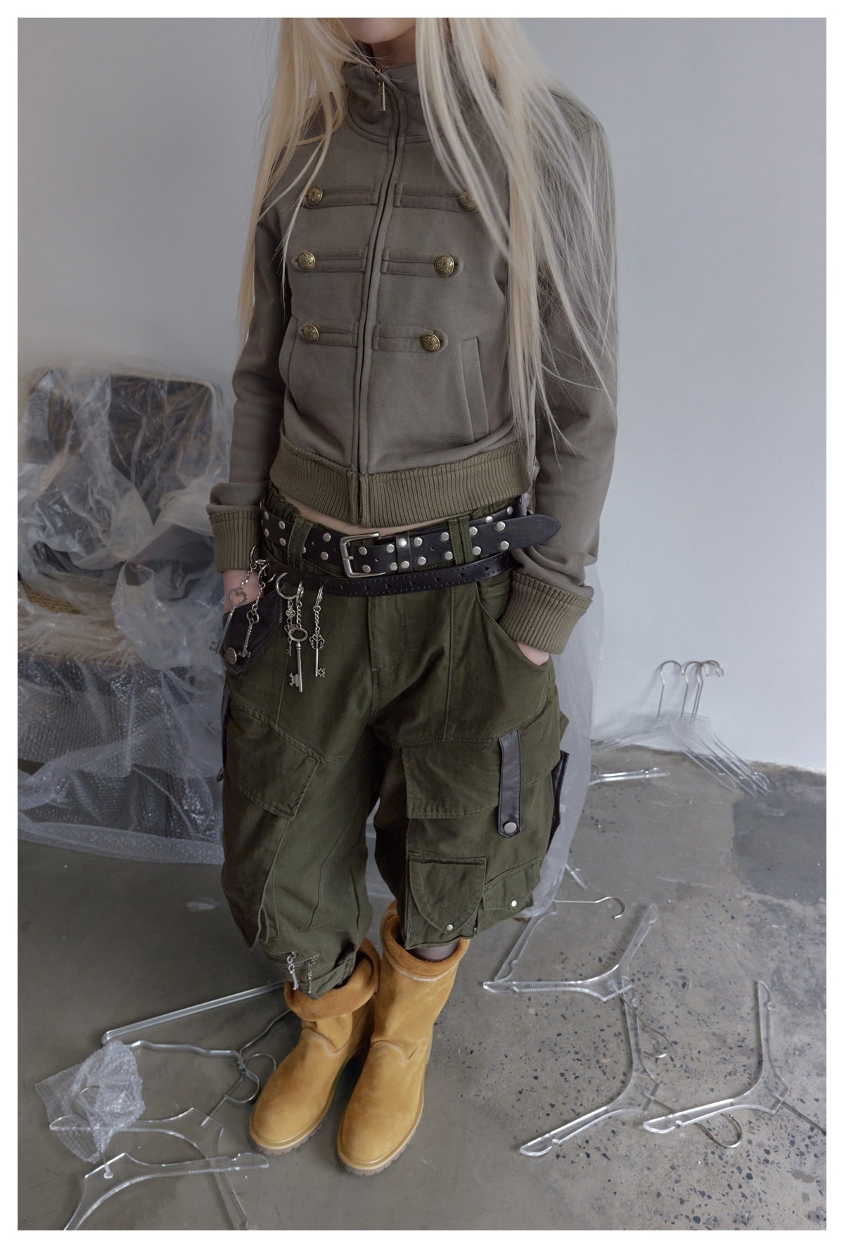 Model wearing a taupe cropped jacket with brass crest buttons and ribbed trim, olive cargo pants, and tan boots.