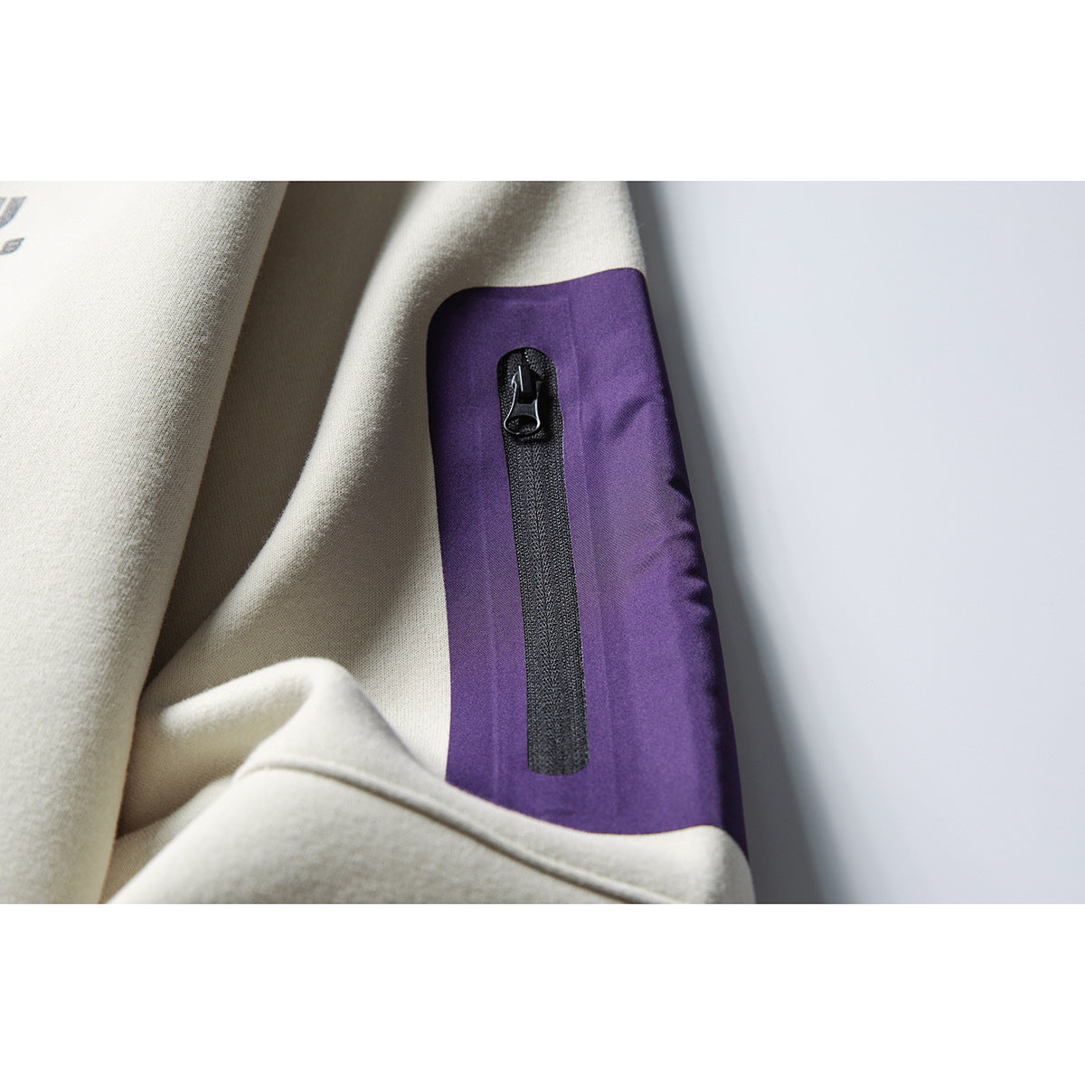 Close-up of a vertical black zipper pocket on a purple sleeve panel of a light beige sweatshirt.
