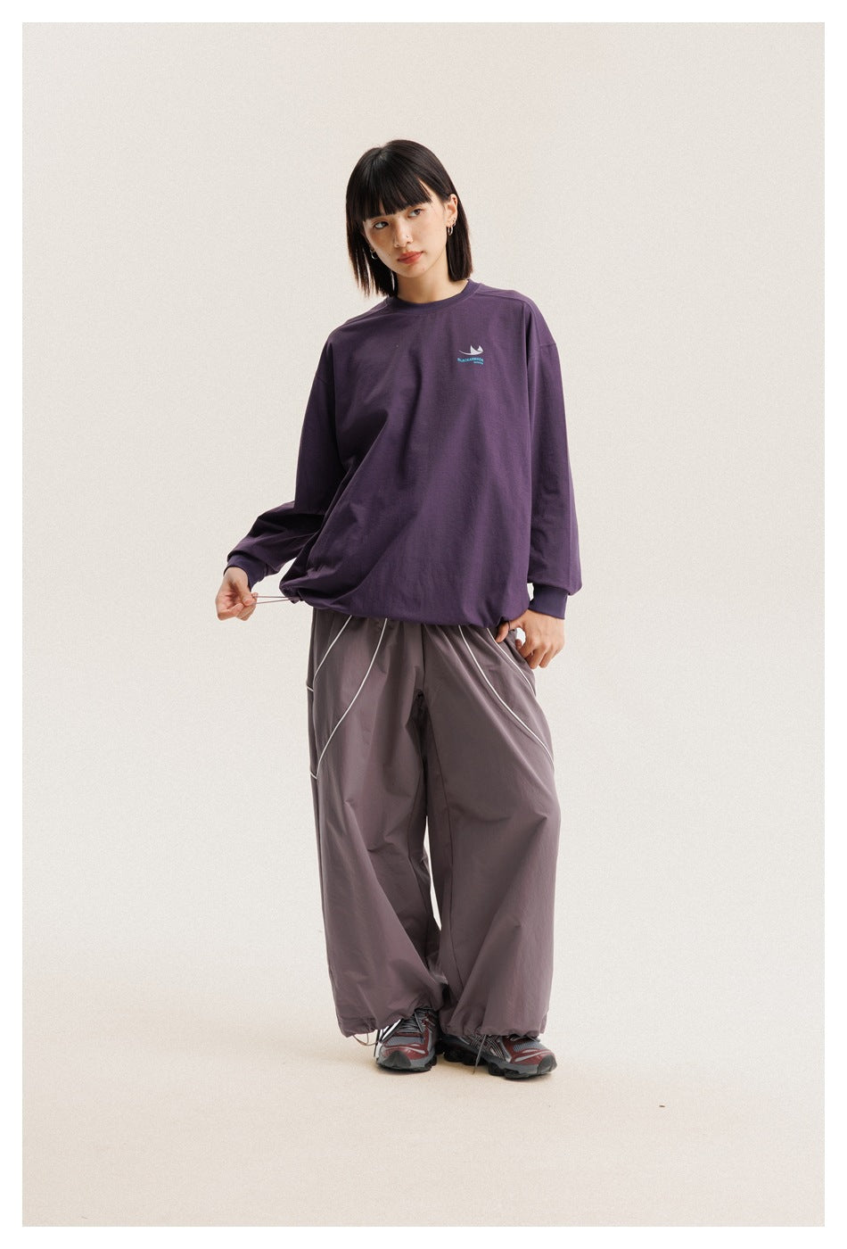 Model pulls the hem of an oversized purple long-sleeve shirt, paired with loose gray-mauve pants.