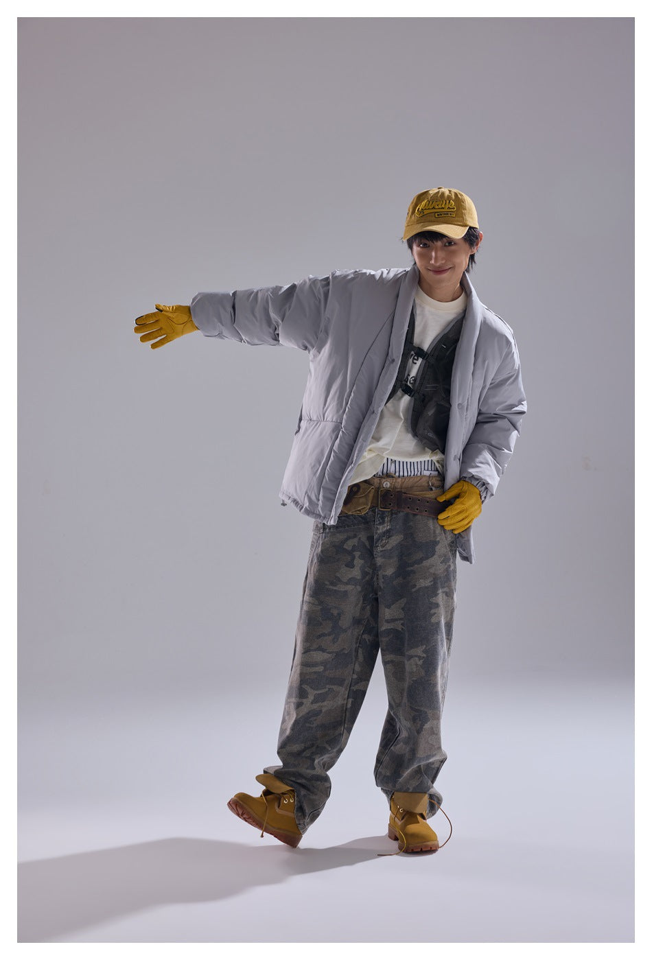 Model wearing the gray padded jacket open over a white tee with camouflage pants, yellow cap and gloves