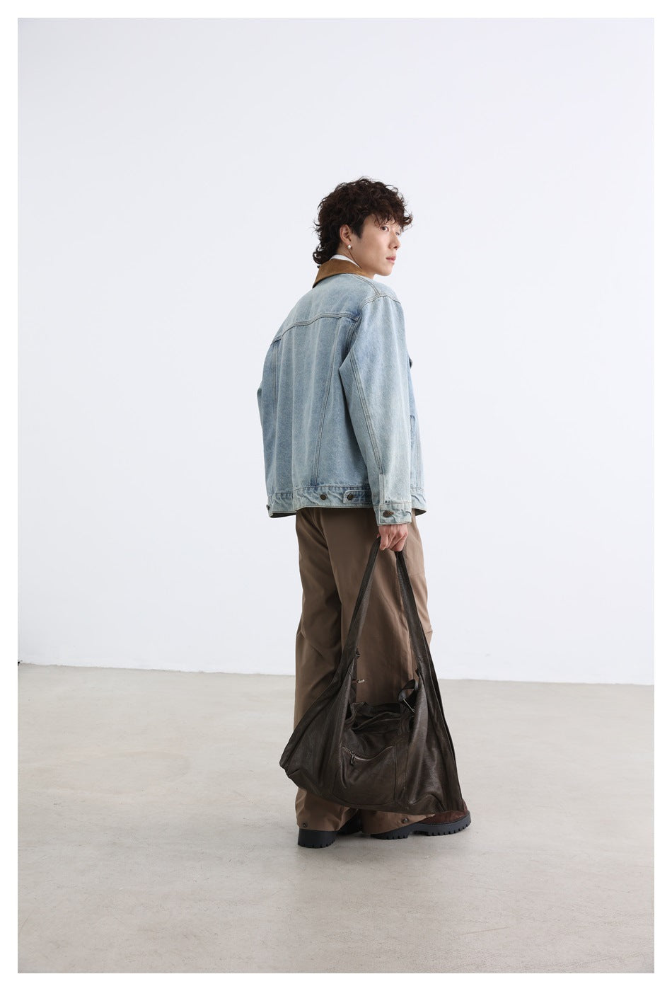 Model turned away to show the back of a light-wash denim jacket with a brown collar; carrying a dark tote bag.