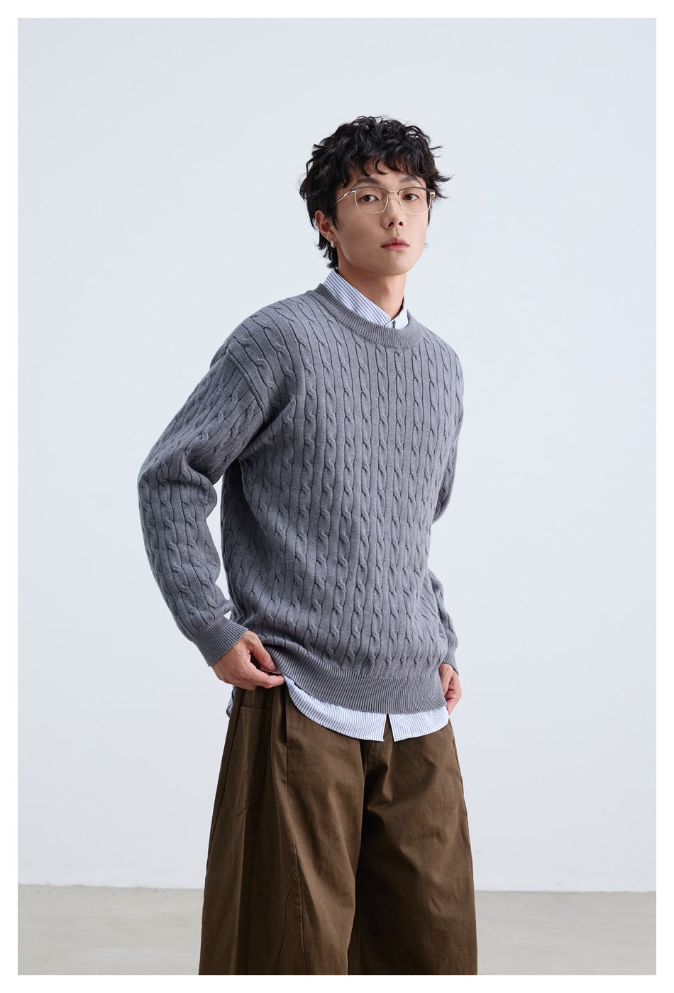 Model wearing a gray cable-knit crewneck over a striped shirt with brown trousers.