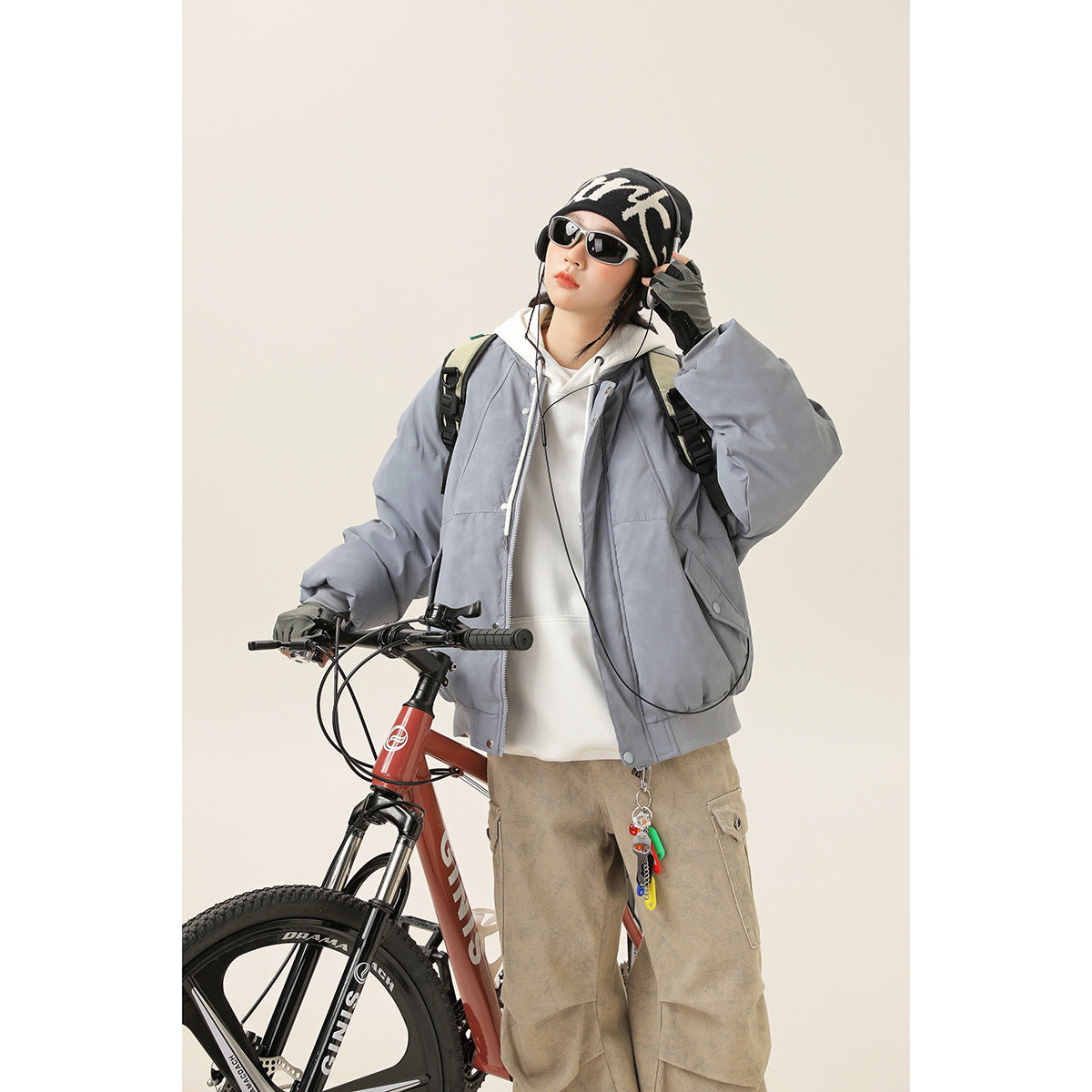 Model wearing an oversized blue-gray bomber jacket over a white hoodie, posed beside a bicycle.