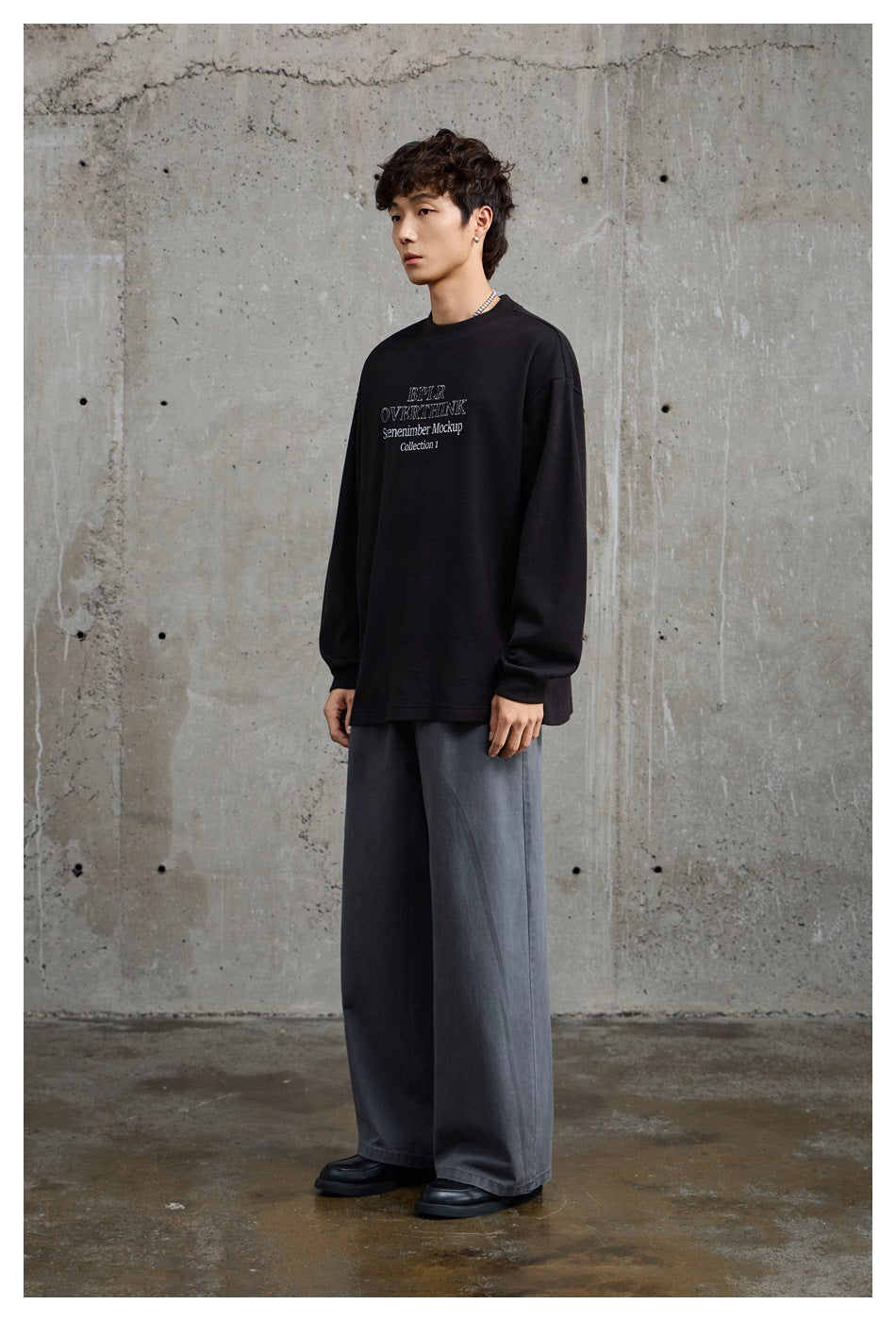 Model in a black oversized crewneck with white chest lettering, shown at an angle with loose gray trousers.