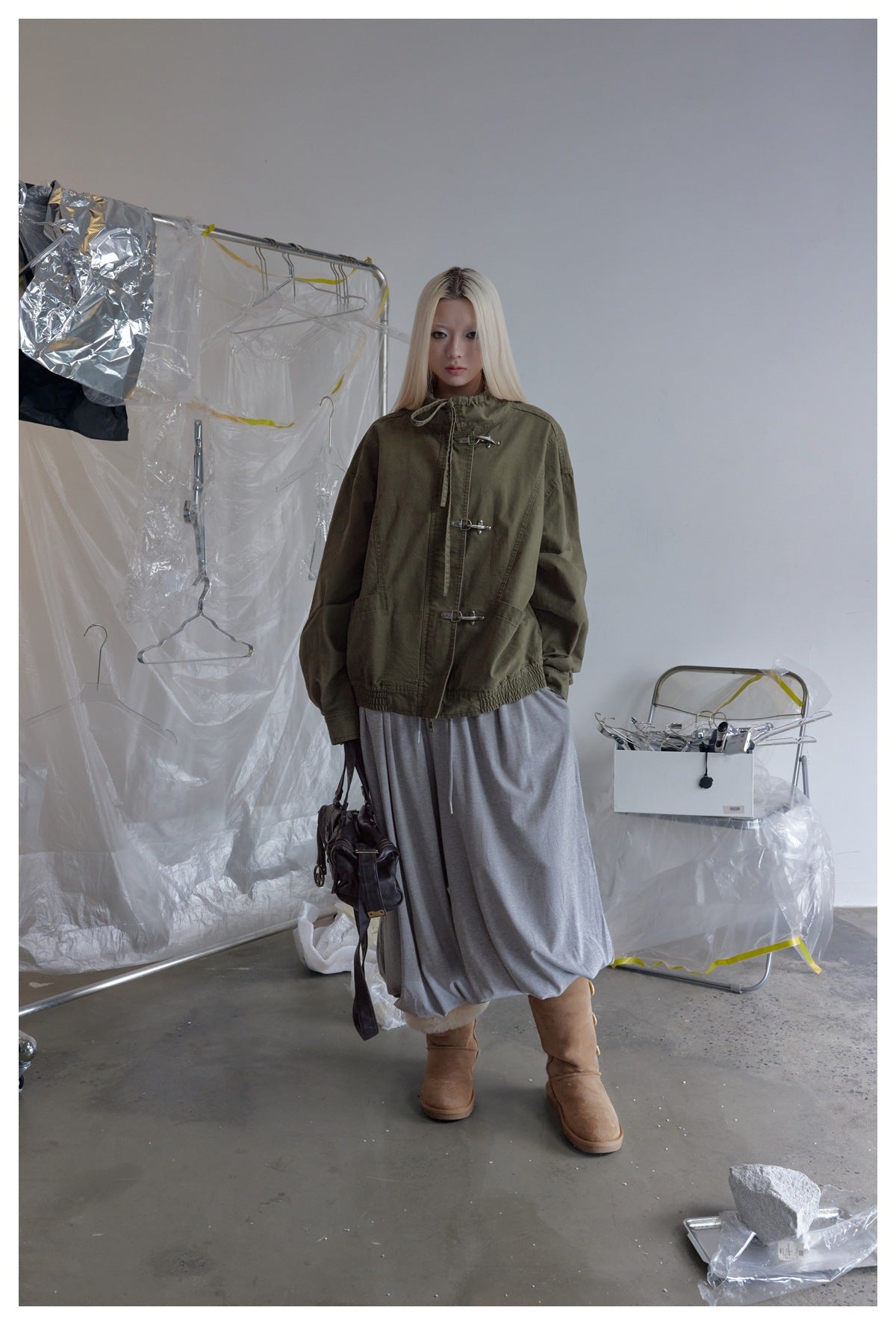 Model wearing the olive green stand-collar jacket fastened with metal hooks, gray skirt and tan boots