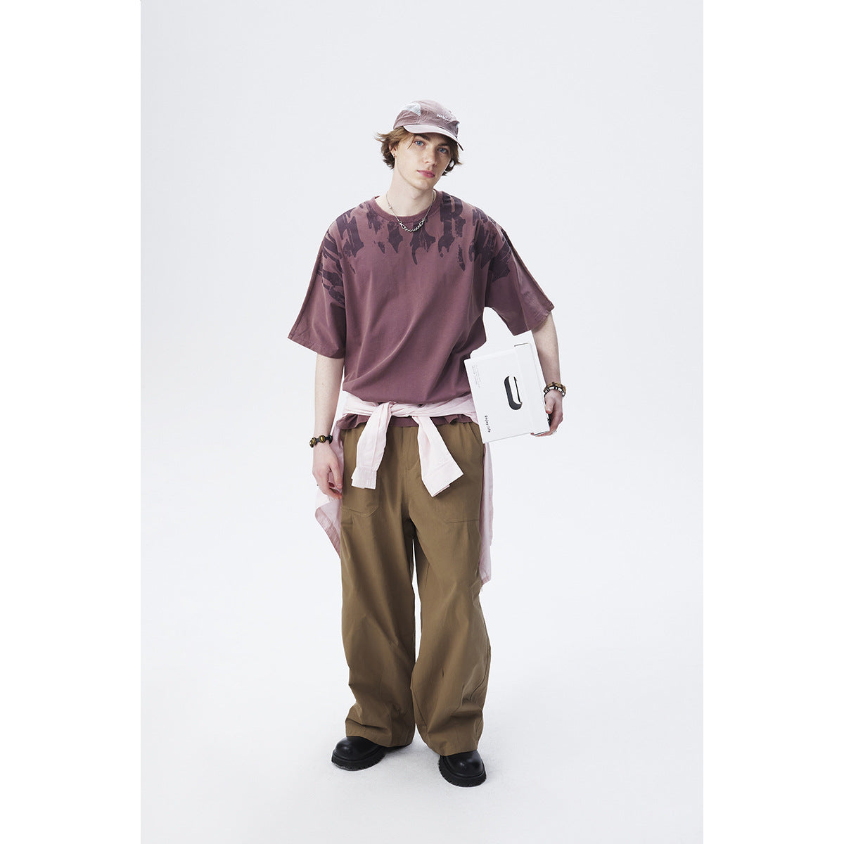 Model wearing a mauve oversized T-shirt with dark abstract shoulder print and loose brown trousers.