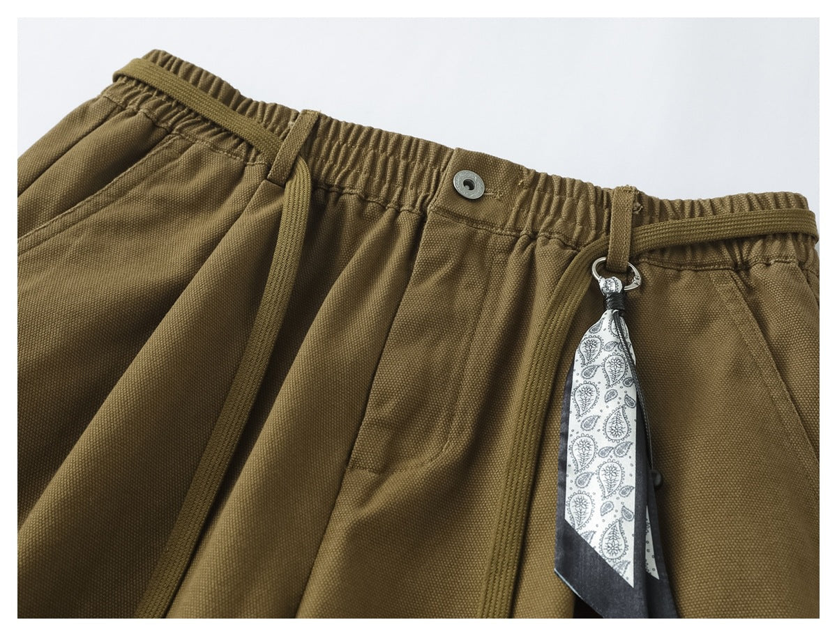 Close-up of olive-brown trousers with an elastic drawstring waist, front pockets and a paisley-patterned hanging accessory.