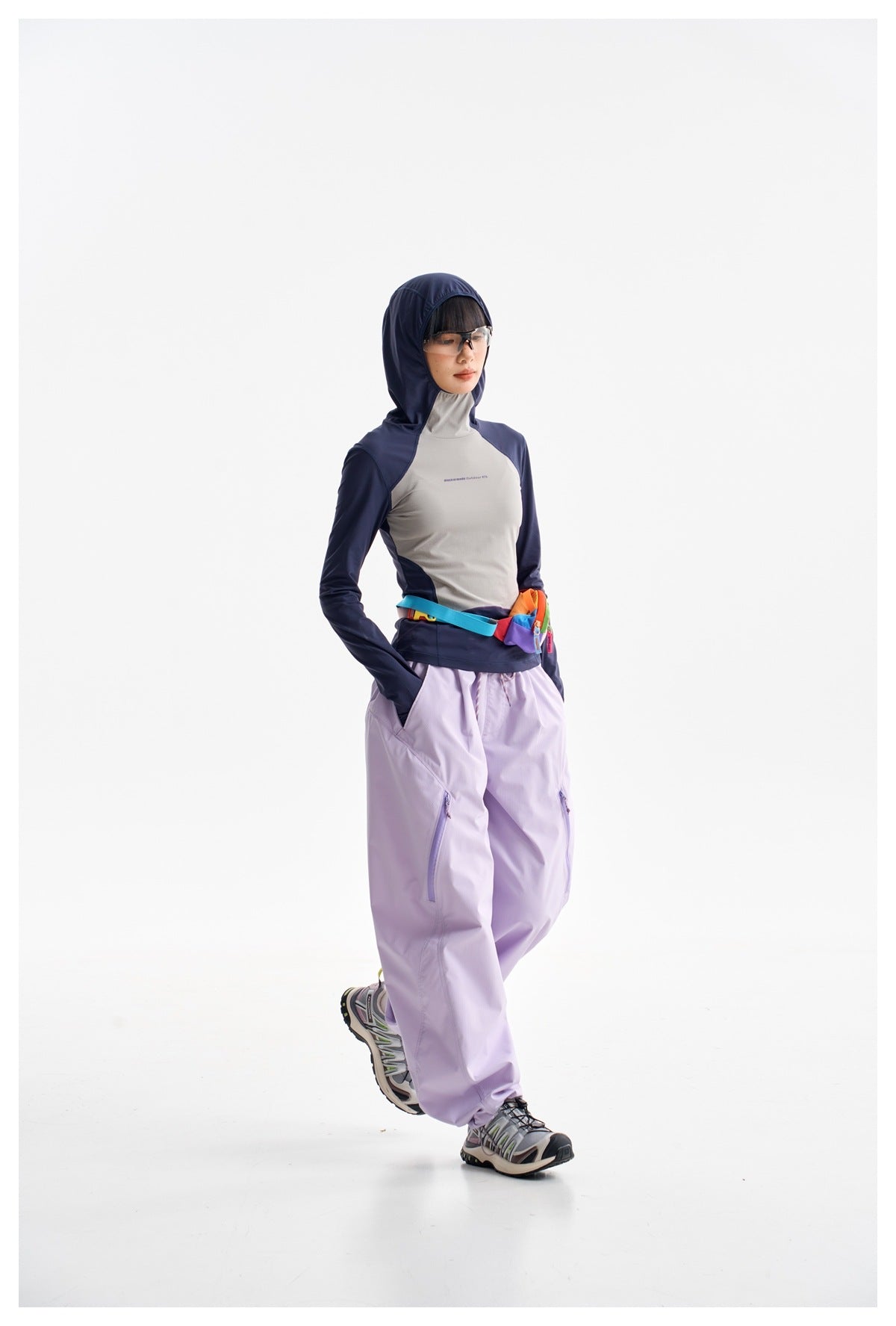 Model in a navy and gray hooded top and loose lavender pants, shown at an angle with hands in pockets.